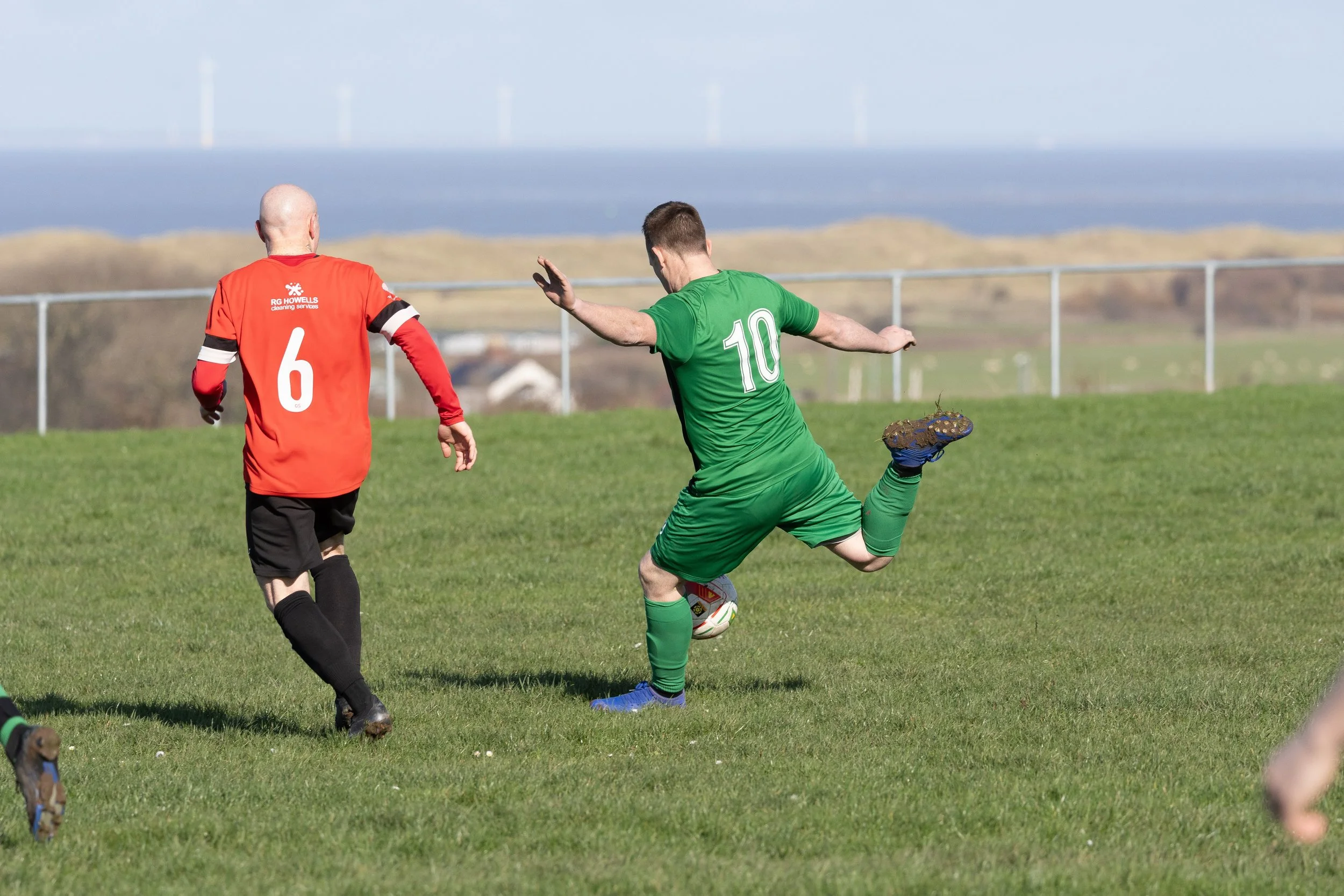 A soccer player in a green uniform with the number 10 on his back is kicking a ball on the field while an opponent in a red uniform with the number 6 is approaching him from behind.