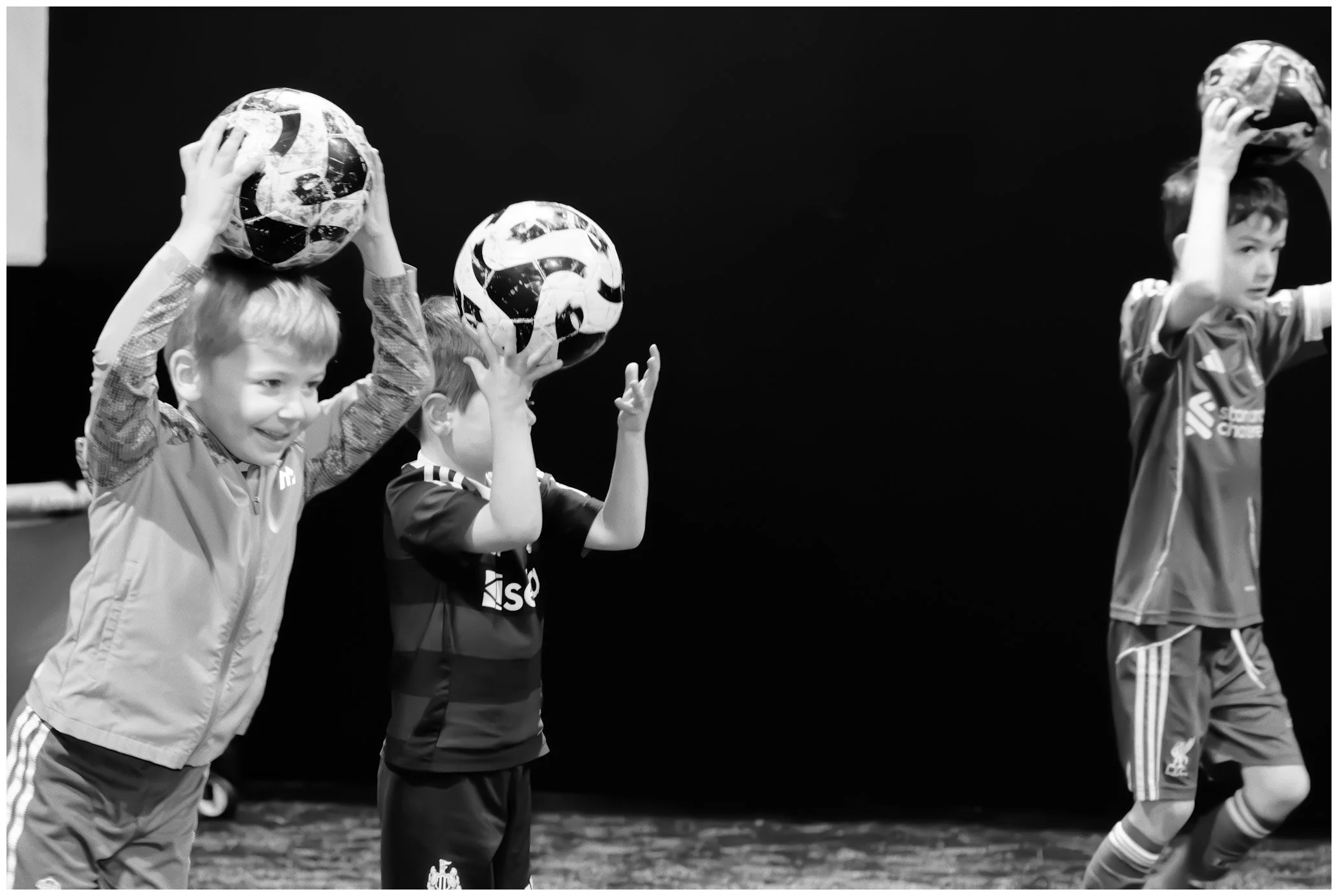 Three young boys standing on a stage holding soccer balls above their heads, dressed in sports uniforms, with a dark background.