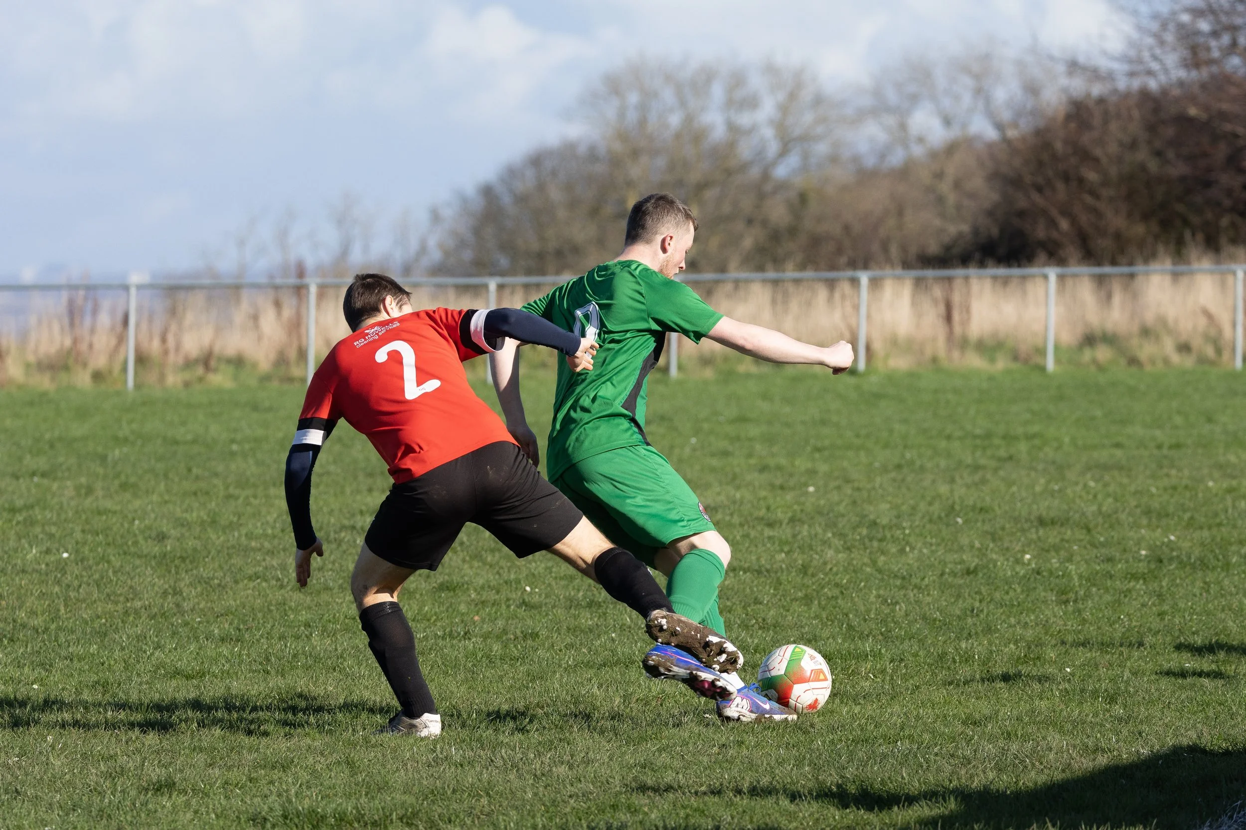 Two soccer players competing for the ball on a grassy field, one wearing a red jersey with the number 2 and black shorts, and the other in a green jersey and shorts, with a background of trees and a blue sky.