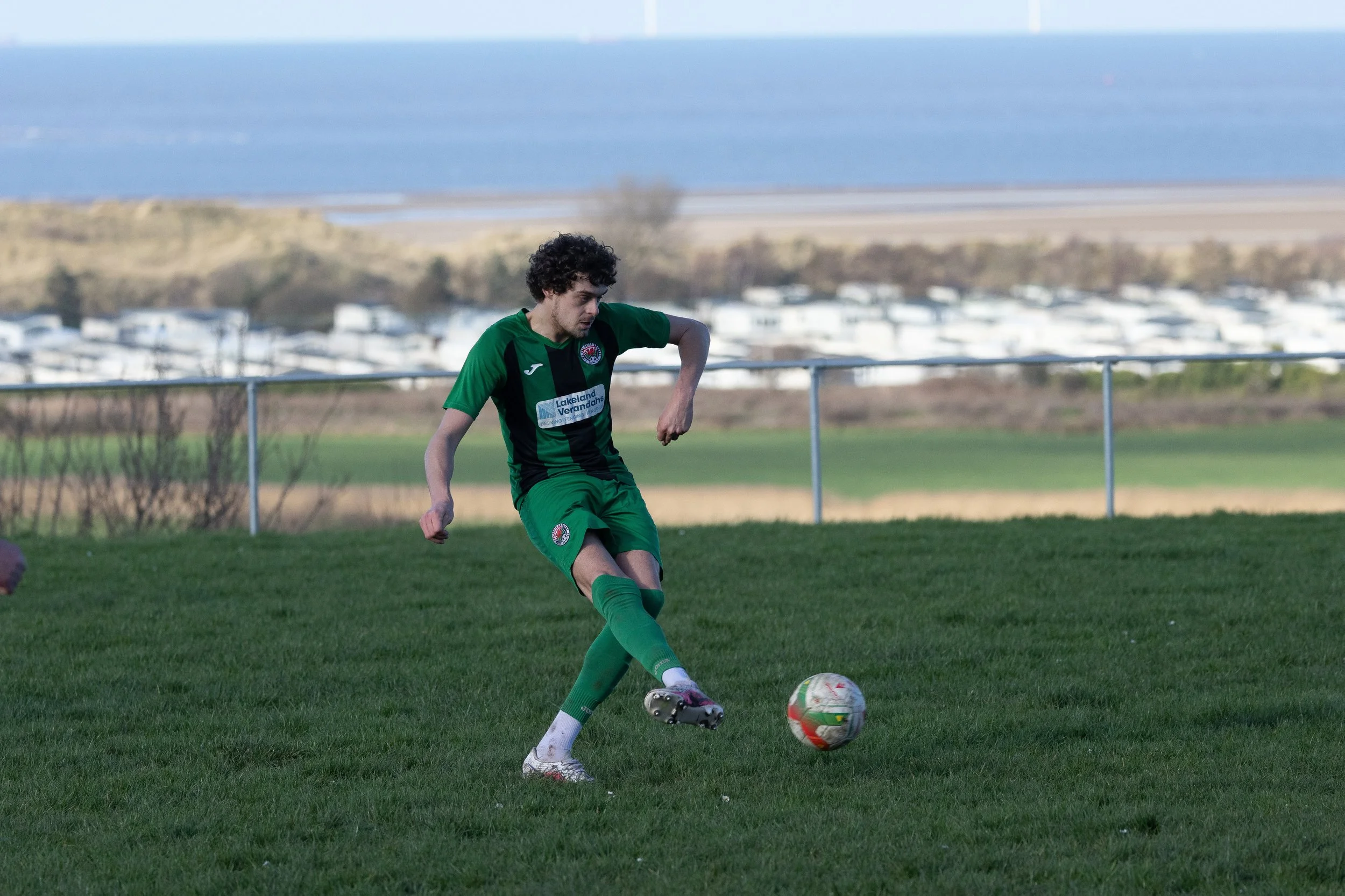 Soccer player in green uniform kicks a ball on a grassy field with a coastal landscape in the background.