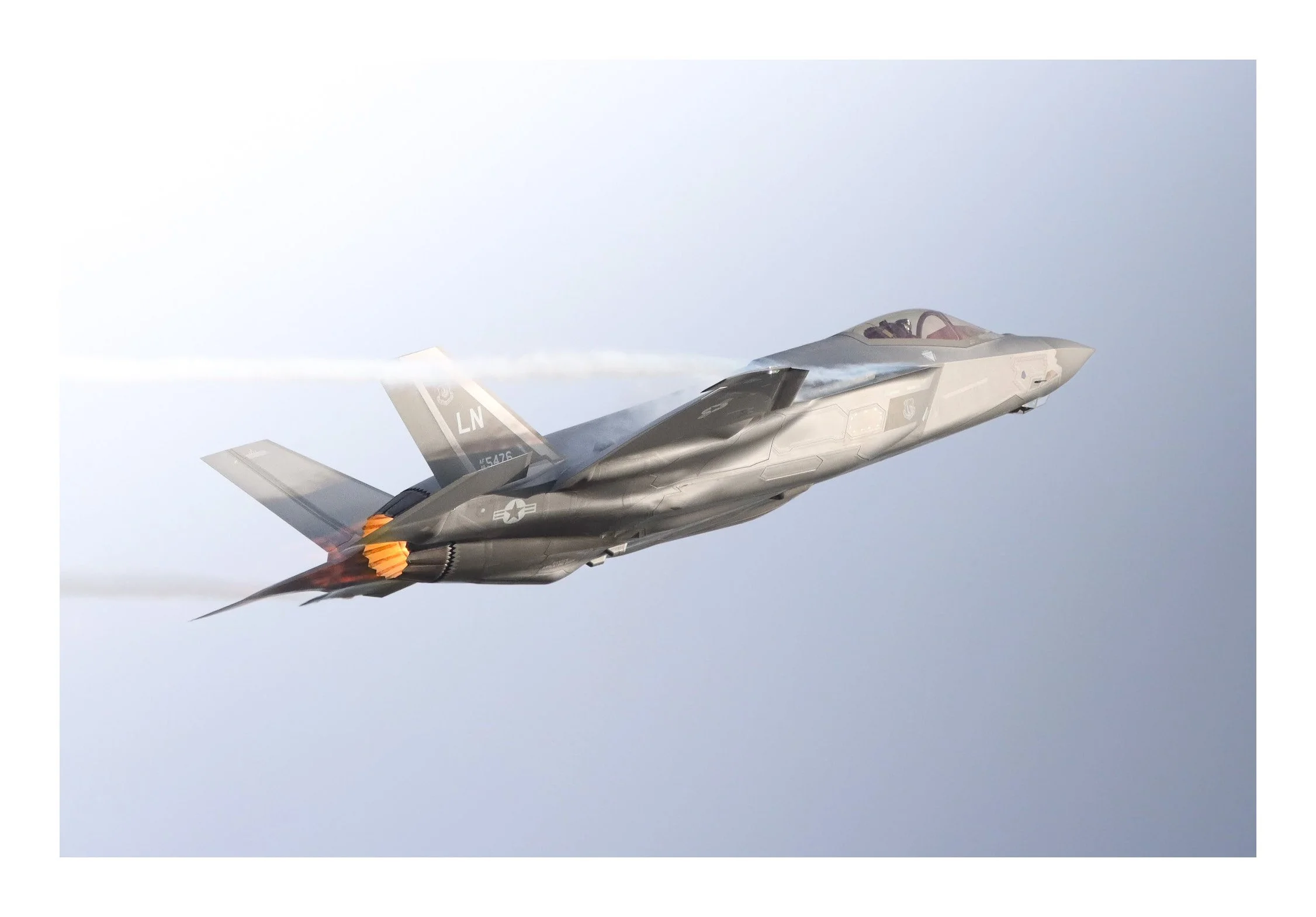 A fighter jet aircraft in flight, with afterburners engaged, emitting a trail of white smoke against a cloudy sky.