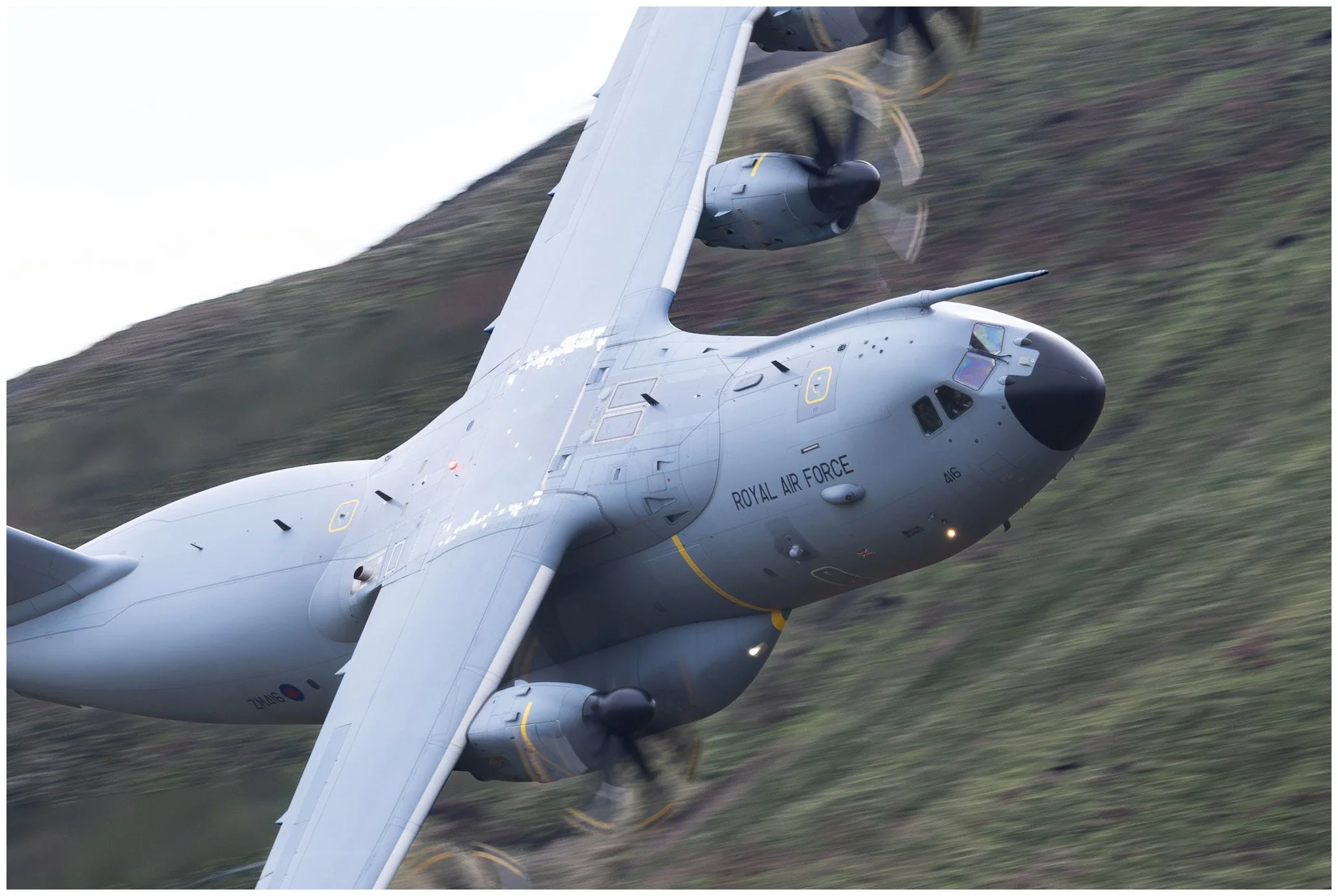 A Royal Air Force military transport aircraft flying at an angle over a green landscape, with visible propellers and a black nose cone.