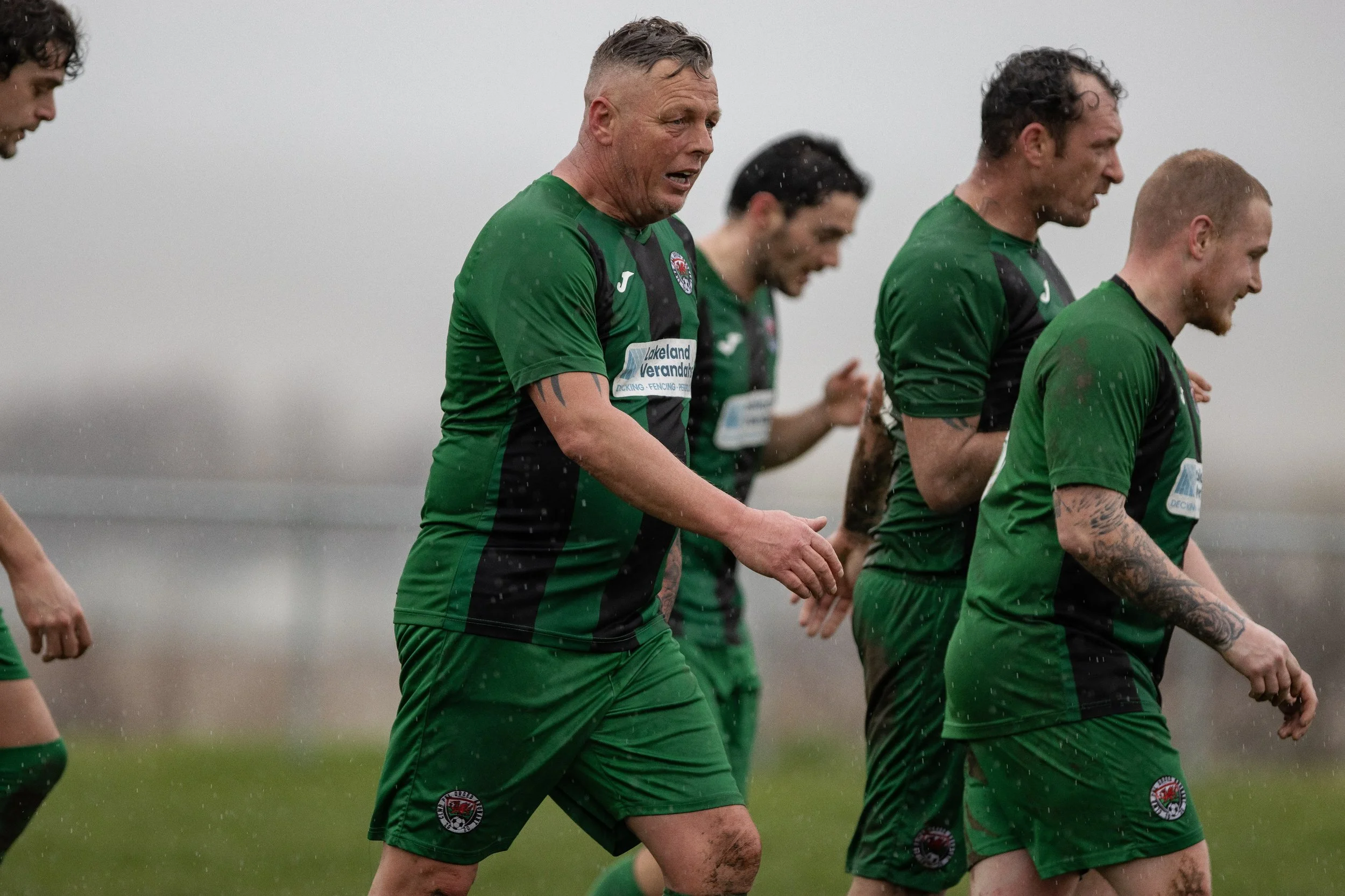 Group of soccer players in green uniforms walking on a muddy grassy field in rainy weather.