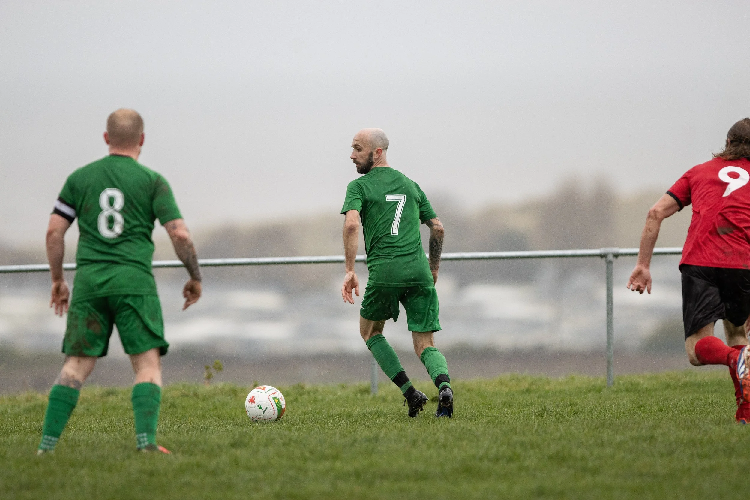 Soccer players in green and red uniforms playing on a field on a gloomy day with foggy weather.