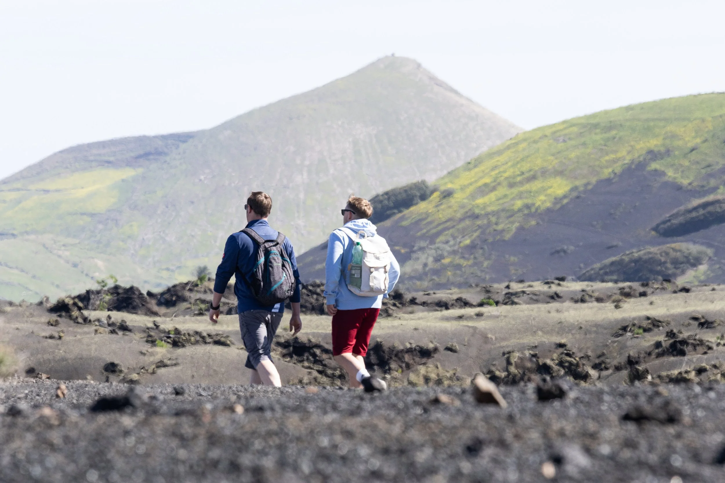 Two hikers walking on volcanic terrain with mountains in the background.