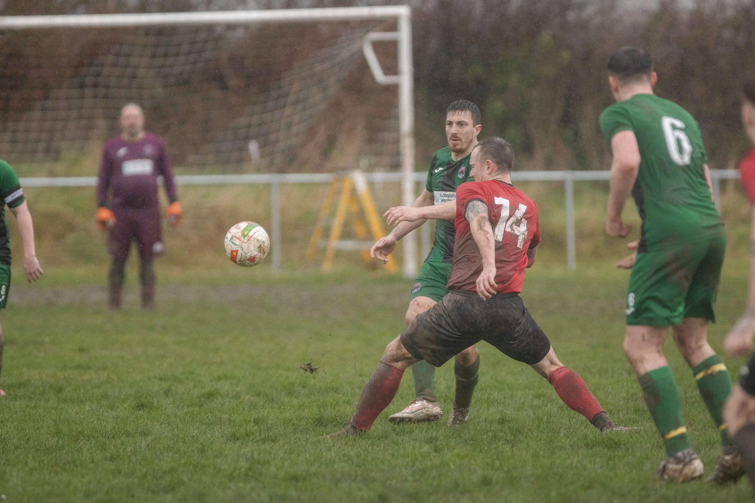 Soccer players struggle for control of the ball on a muddy field during a match. One player in a red and black uniform is falling while trying to reach the ball, with muddy shorts and socks, as others in green jerseys watch or approach.
