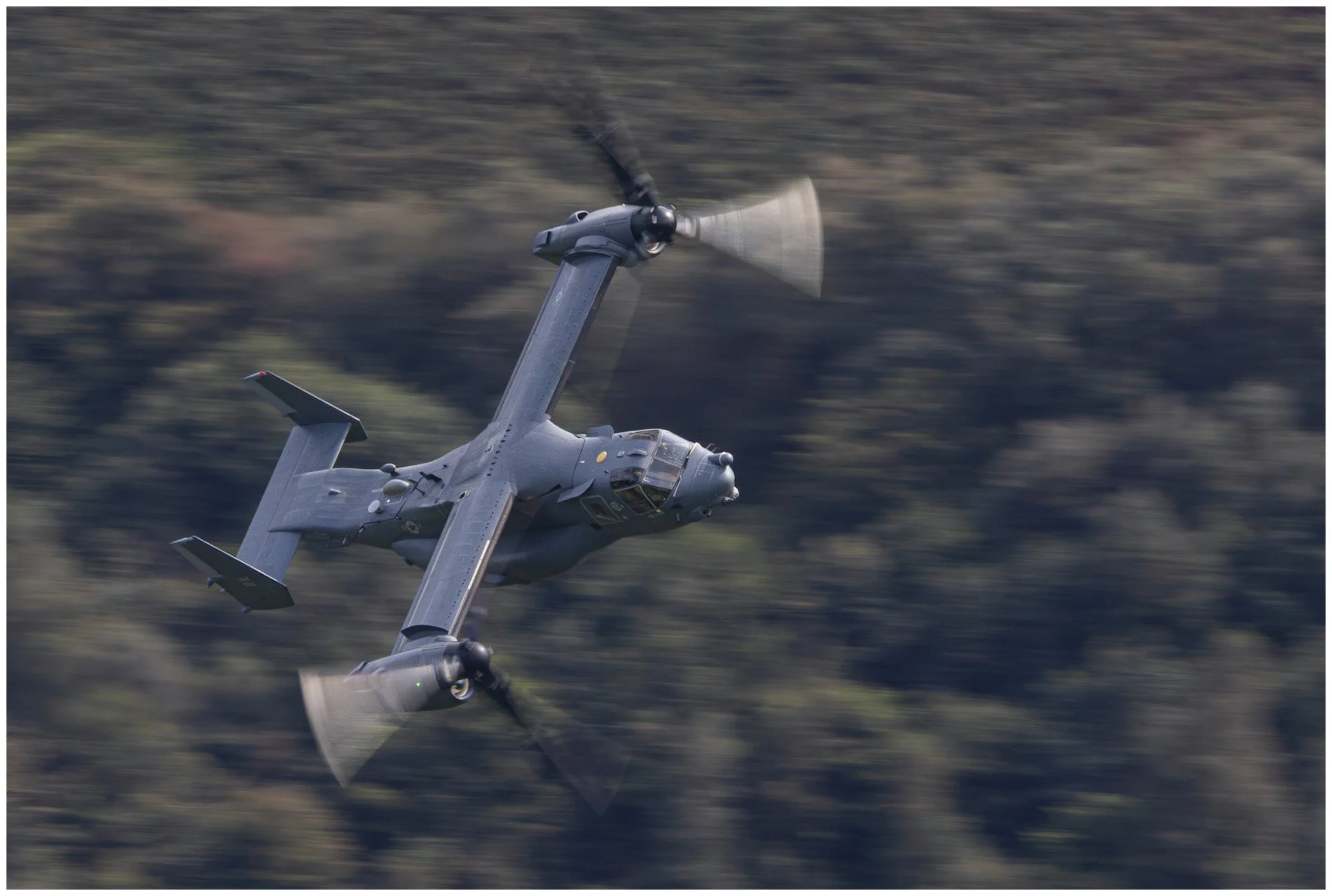 A military tiltrotor aircraft flying at high speed over a forested area with a blurred background.