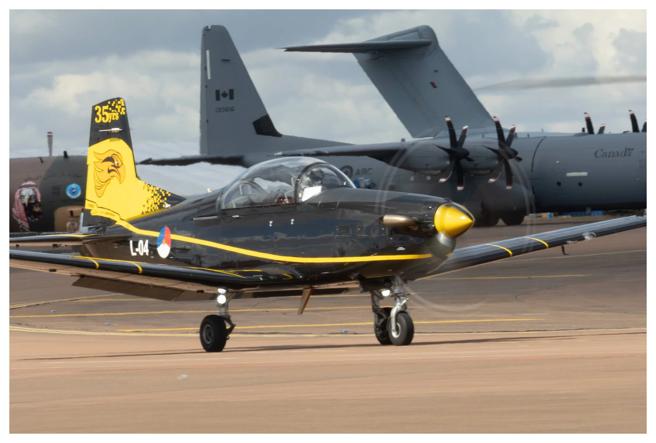 A sleek black and yellow fighter jet taxiing on an airport runway with a large military cargo plane in the background.