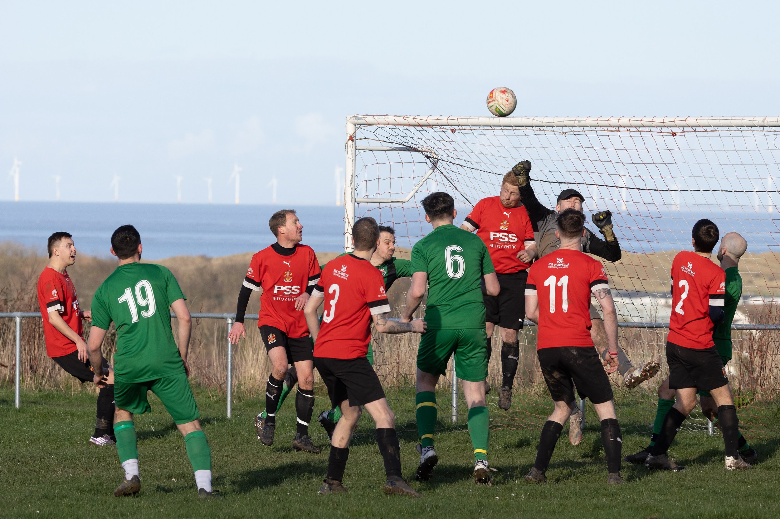 A soccer game in progress with players fighting for a ball near the goal on a grassy field. Some players are wearing red jerseys, while others are in green. The goalkeeper in black is jumping to block the ball in front of the goal. Wind turbines are 