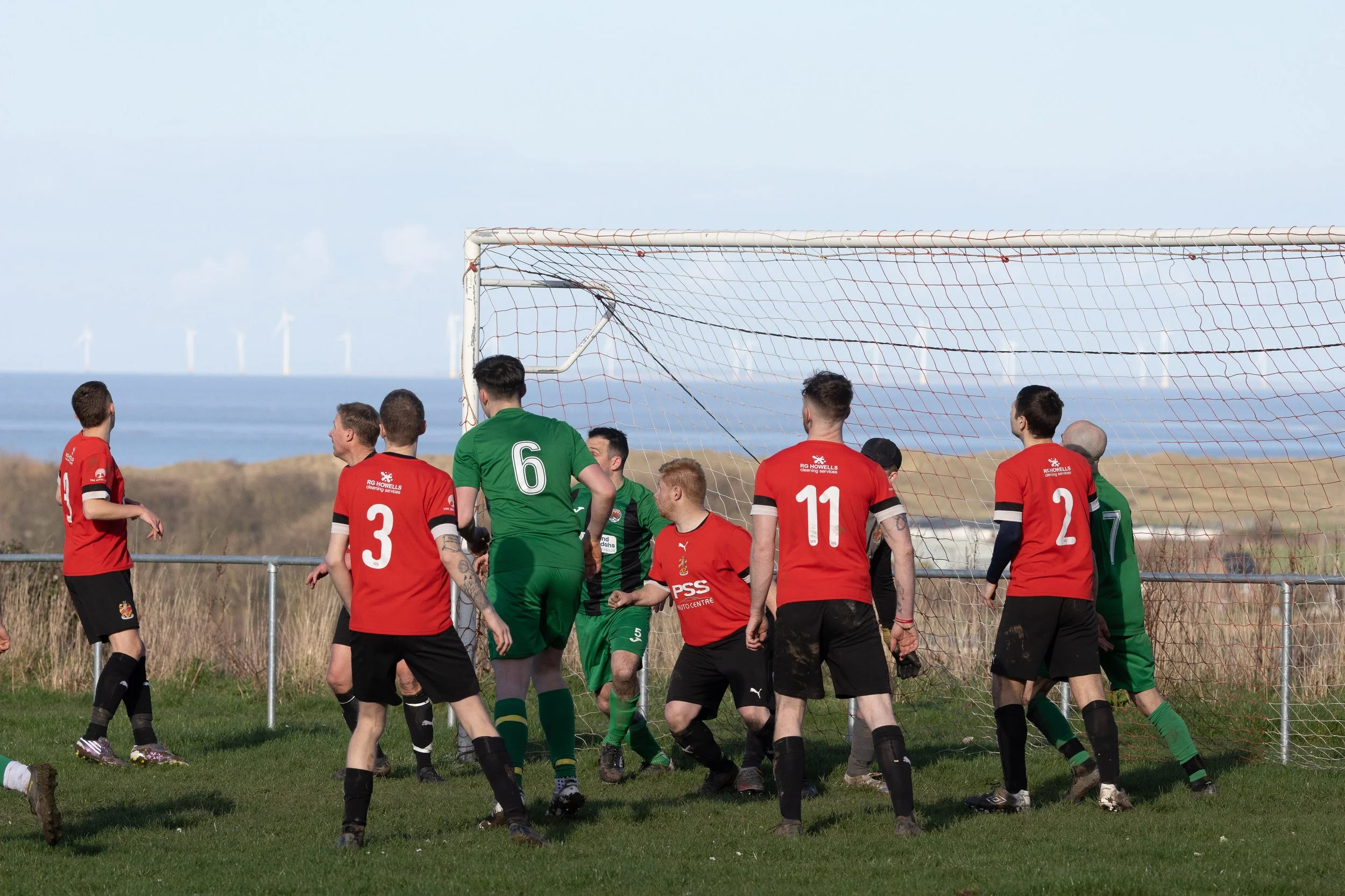 Soccer players battling near the goal during a match, with some players in red and others in green uniforms, on a grassy field near the coast.