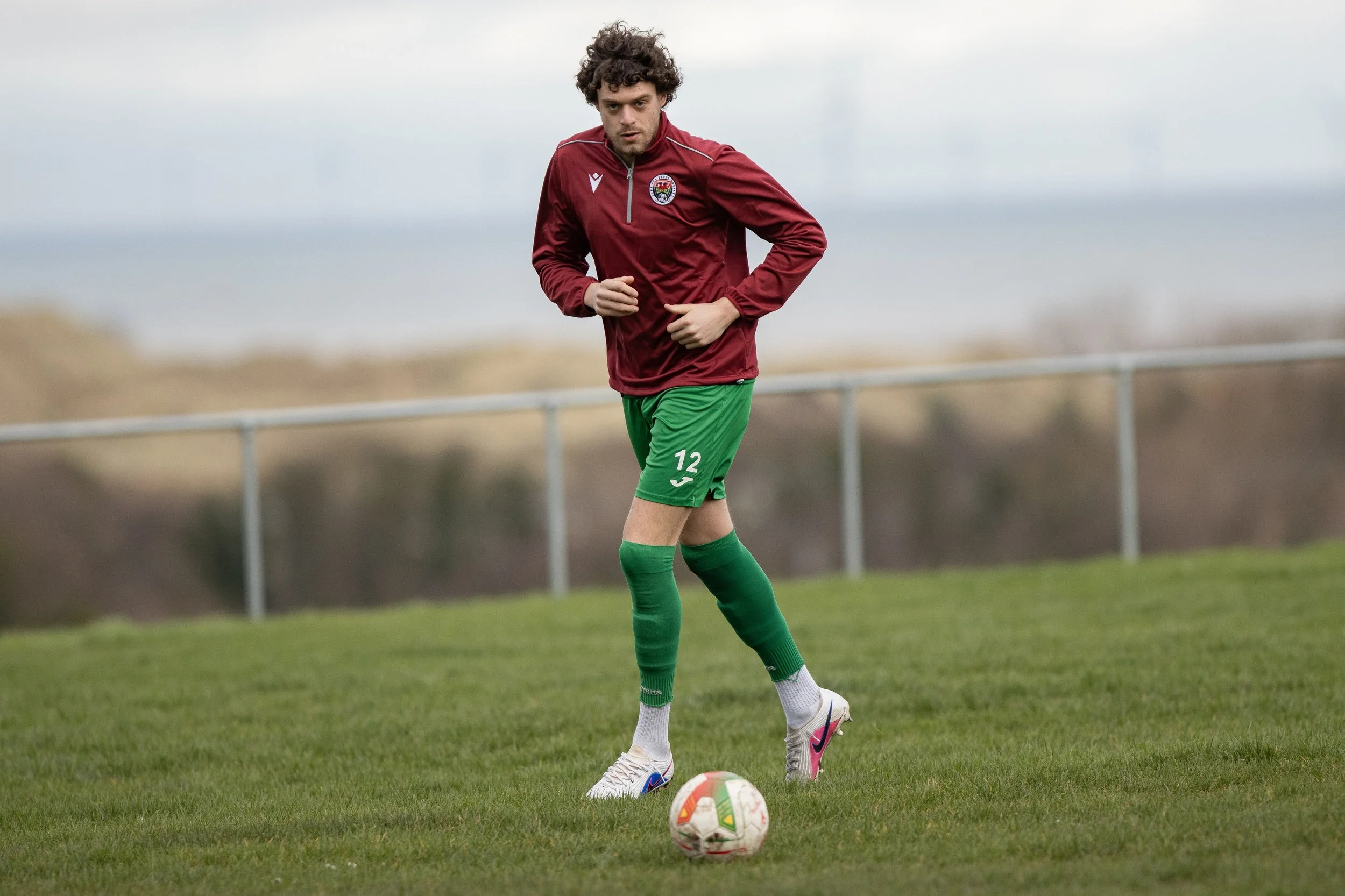 A young man with curly hair in a maroon jacket, green shorts with the number 12, and green socks is on a soccer field, about to kick a colorful soccer ball.