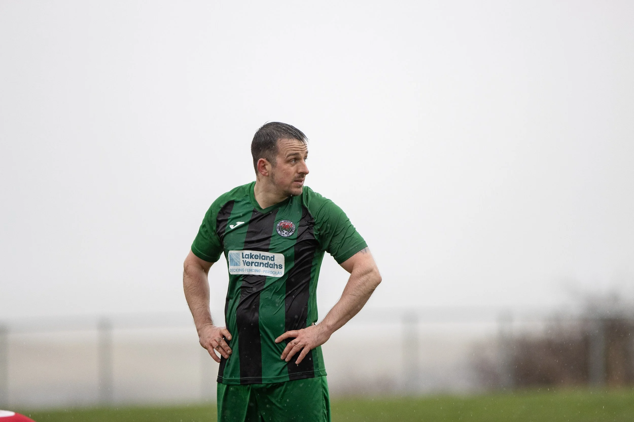 A man in a green and black soccer uniform standing on a field in the rain, with his hands on his hips, looking to the side.