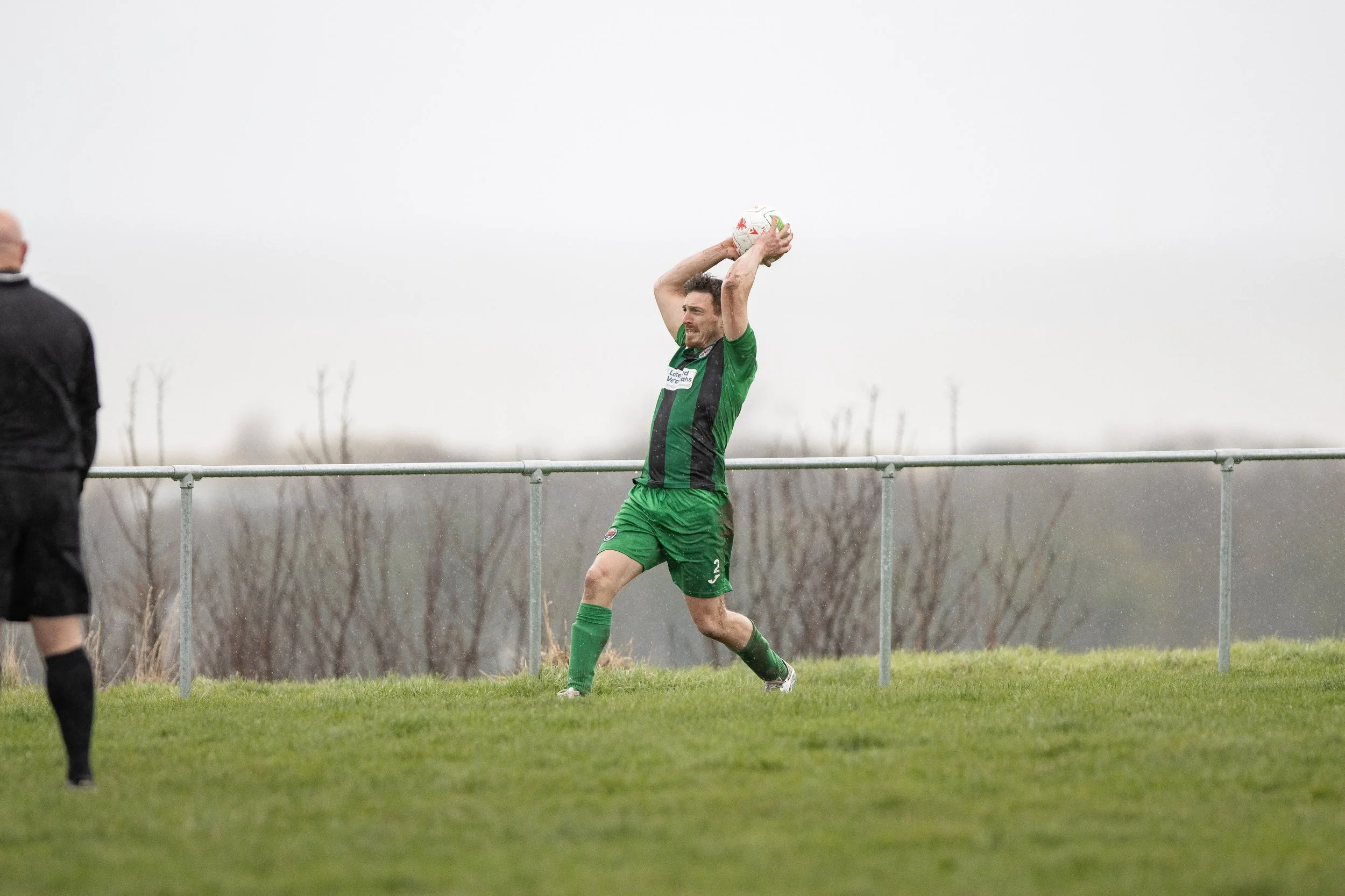 Soccer player in green uniform catching a ball during a game on a grassy field under overcast sky.