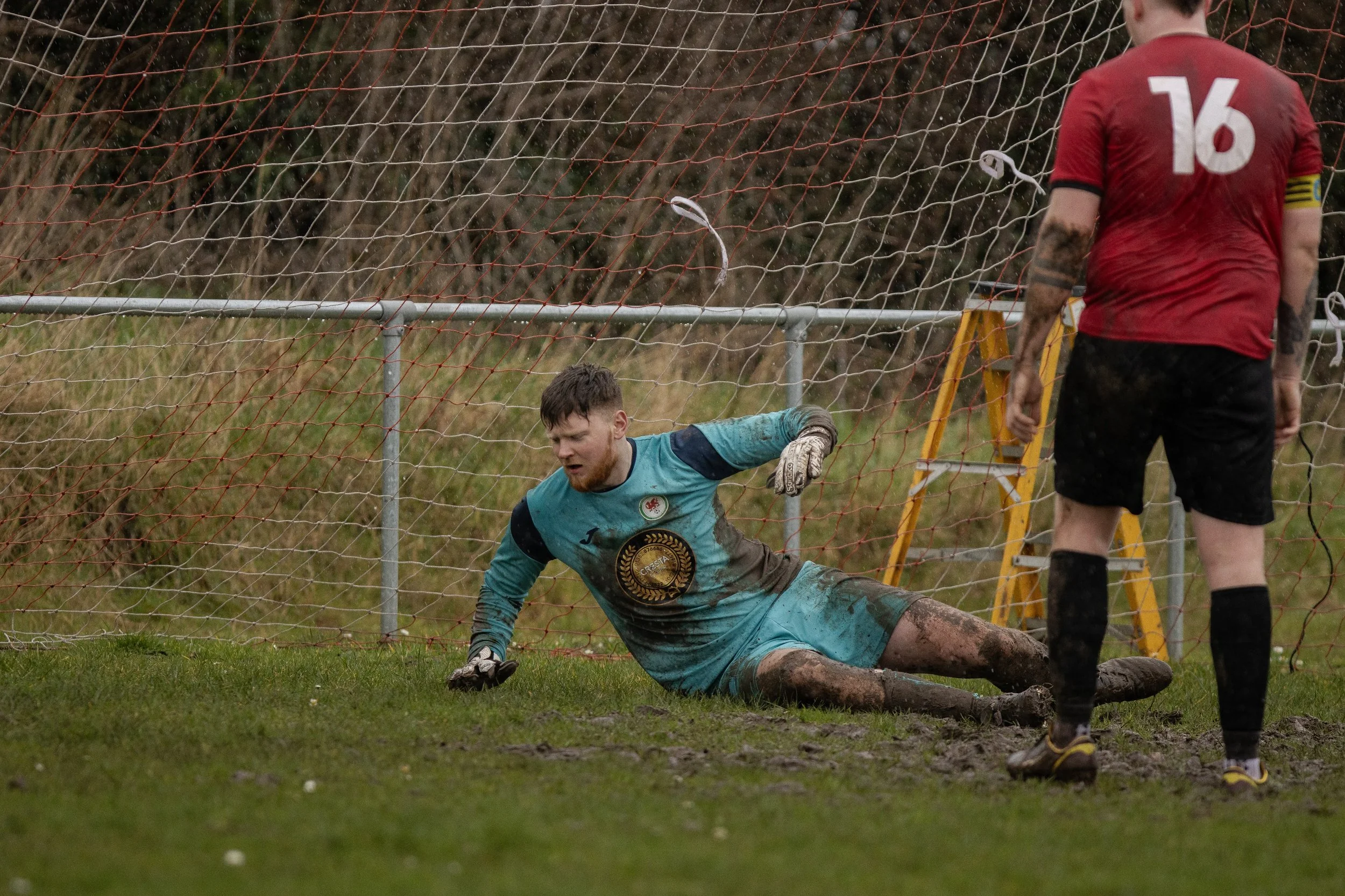 A soccer player in a red jersey with the number 16 standing on a muddy field, watching a goalkeeper in a blue uniform who is on the ground and appears to have just made a save near the goalpost. The scene is outdoors with a muddy ground and a net beh