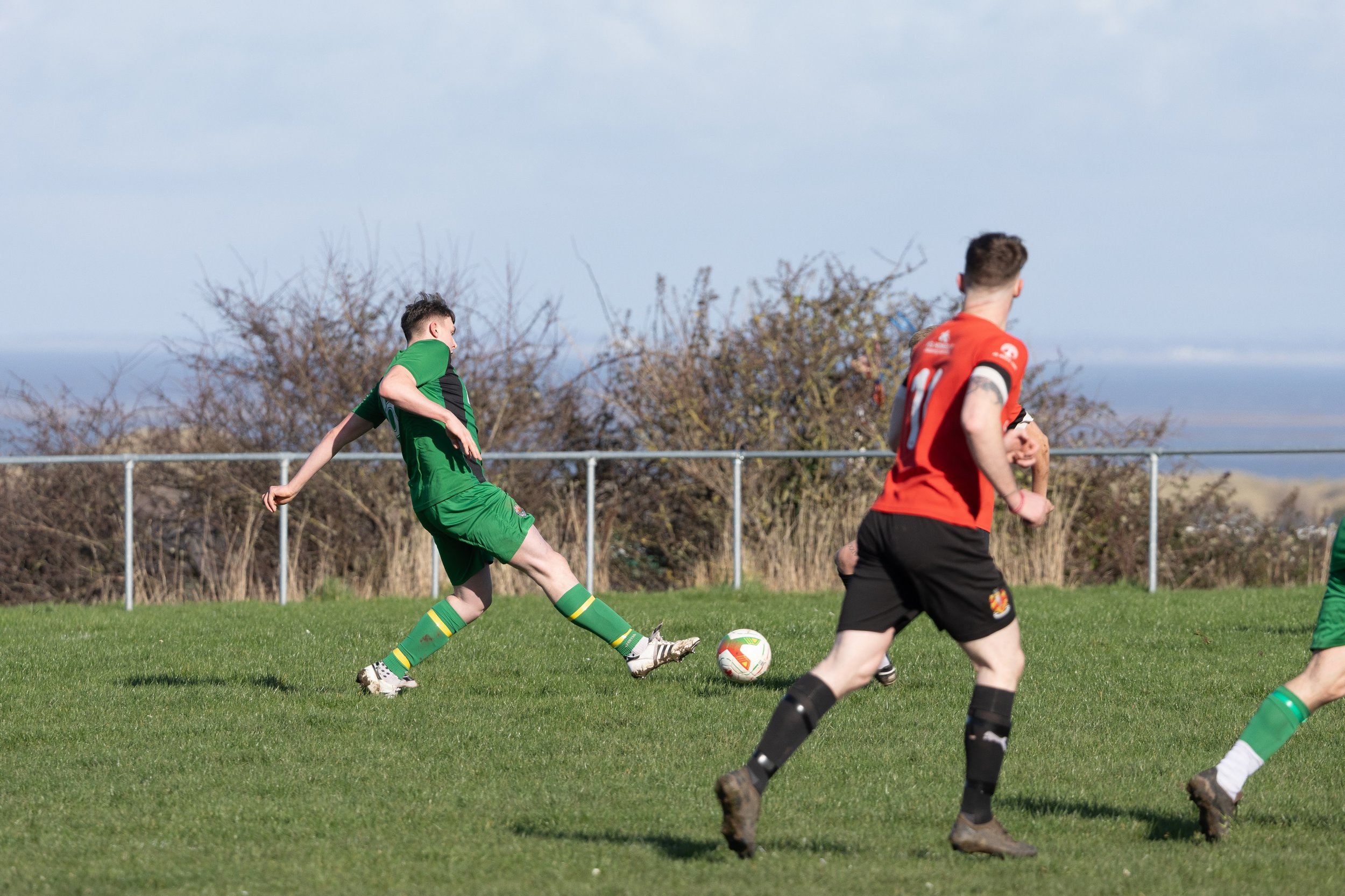Soccer players in green and red jerseys playing on a grass field during daytime, with a blue sky and trees in the background.