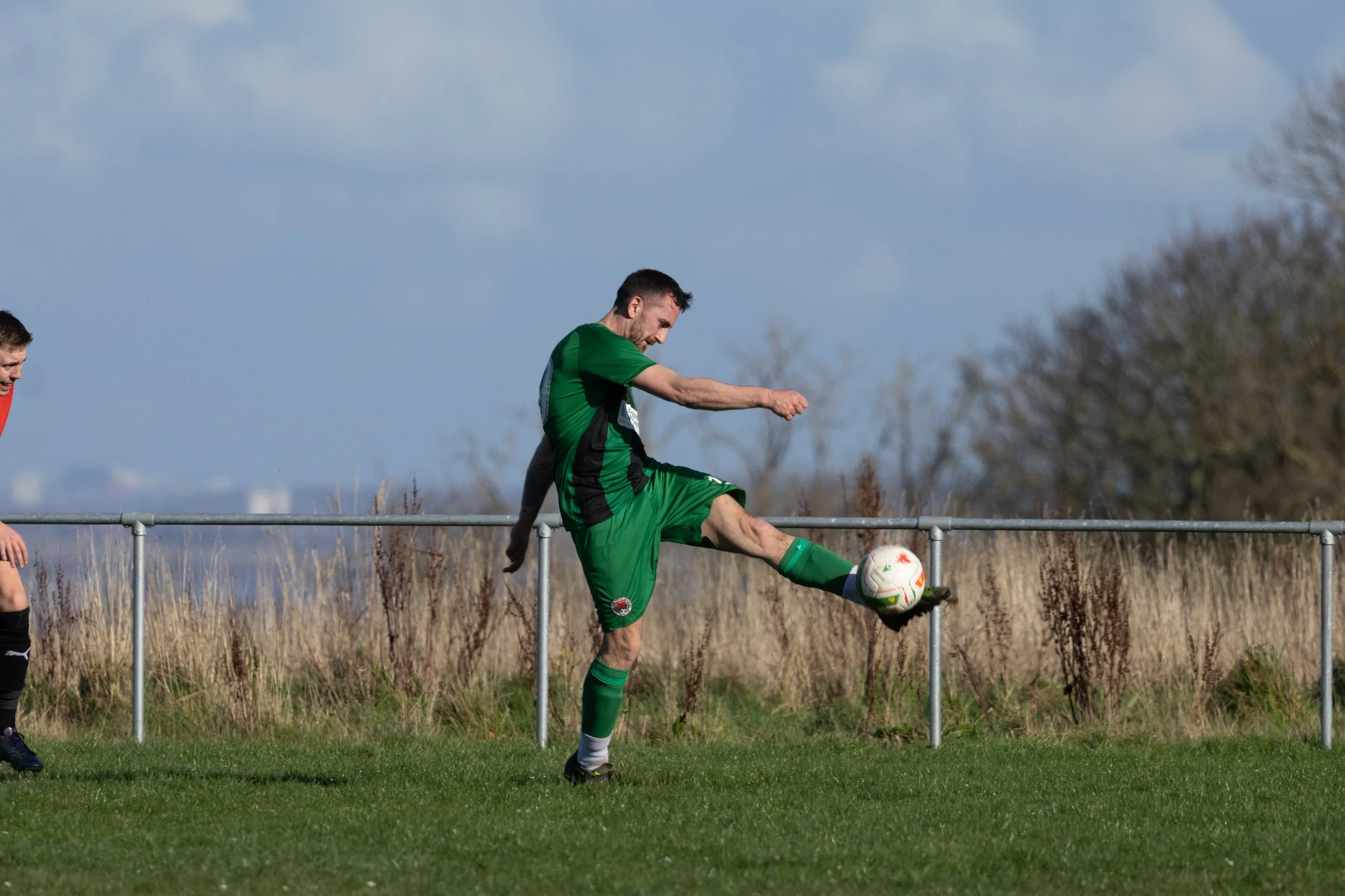 A soccer player in a green uniform kicking a soccer ball on a field with a metal fence and trees in the background.