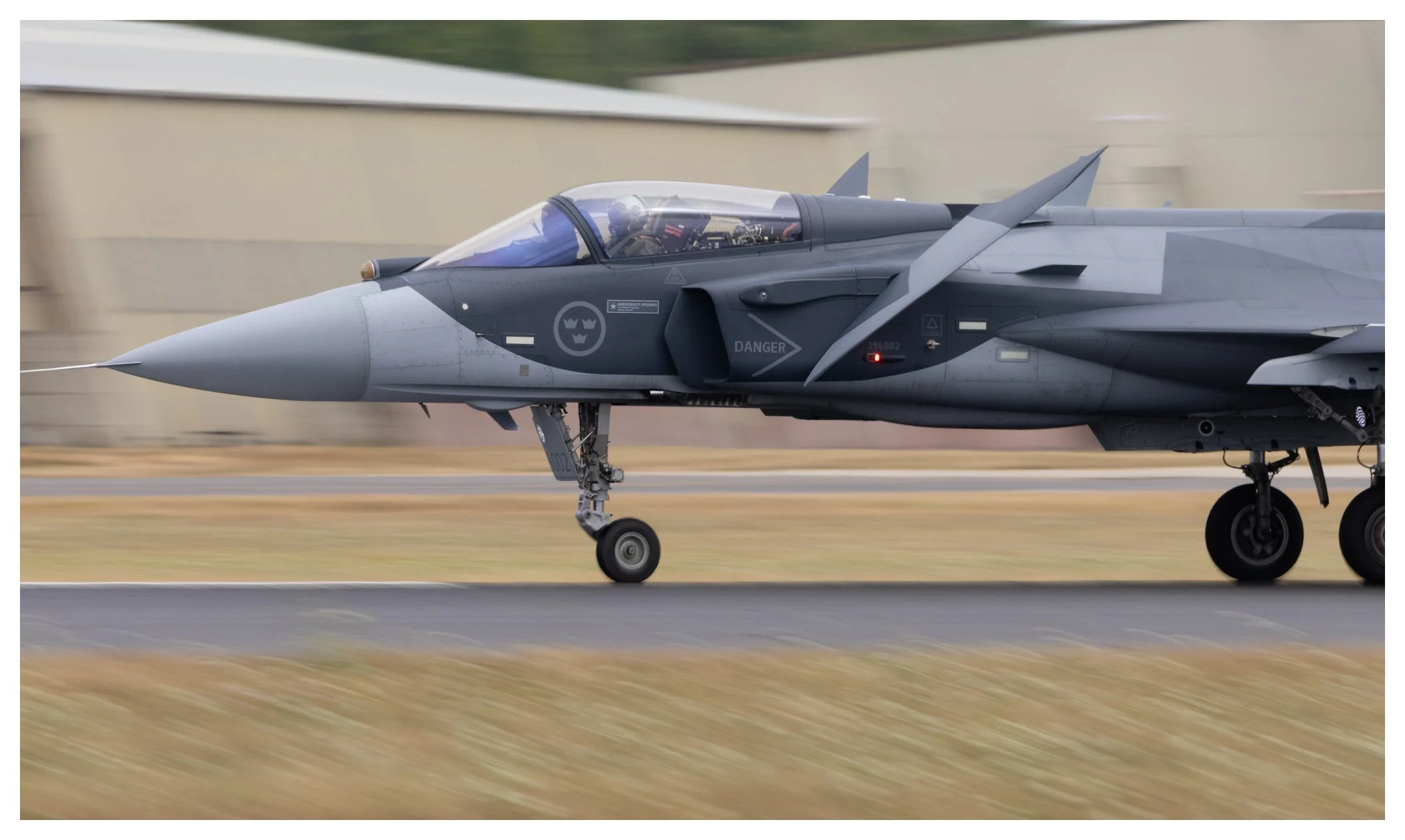 A military fighter jet plane in flight, with wheels on the ground, during takeoff or landing, on a runway with a blurred background.
