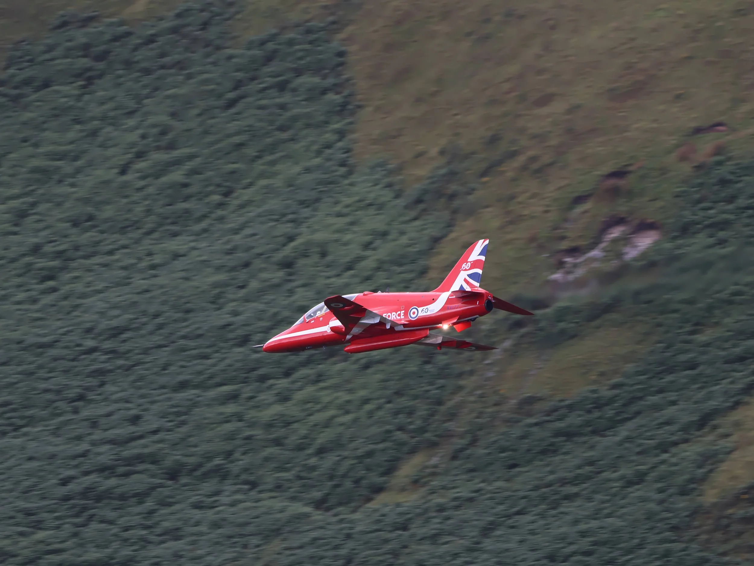 A red aircraft flying over a forested mountain landscape.