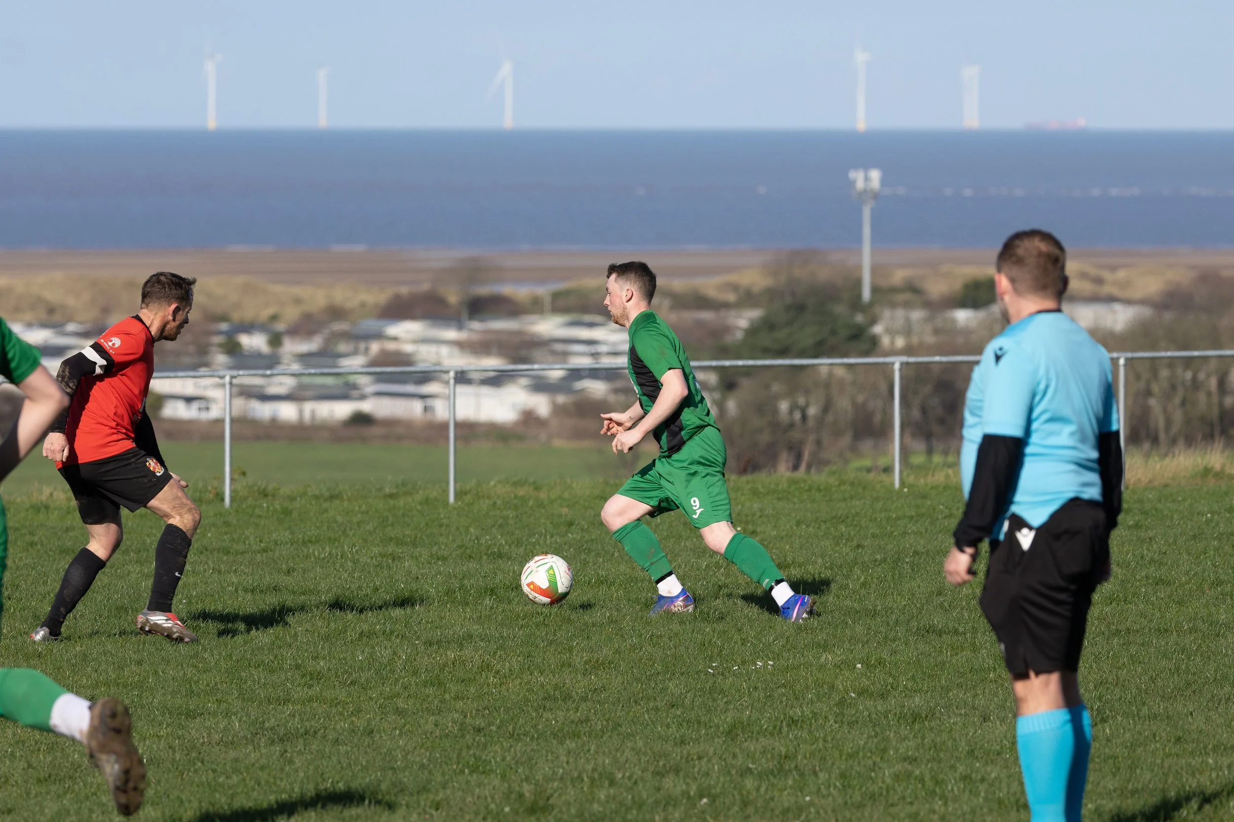 Soccer players in green and red jerseys compete on a grass field with a scenic coastal backdrop, wind turbines on the horizon.