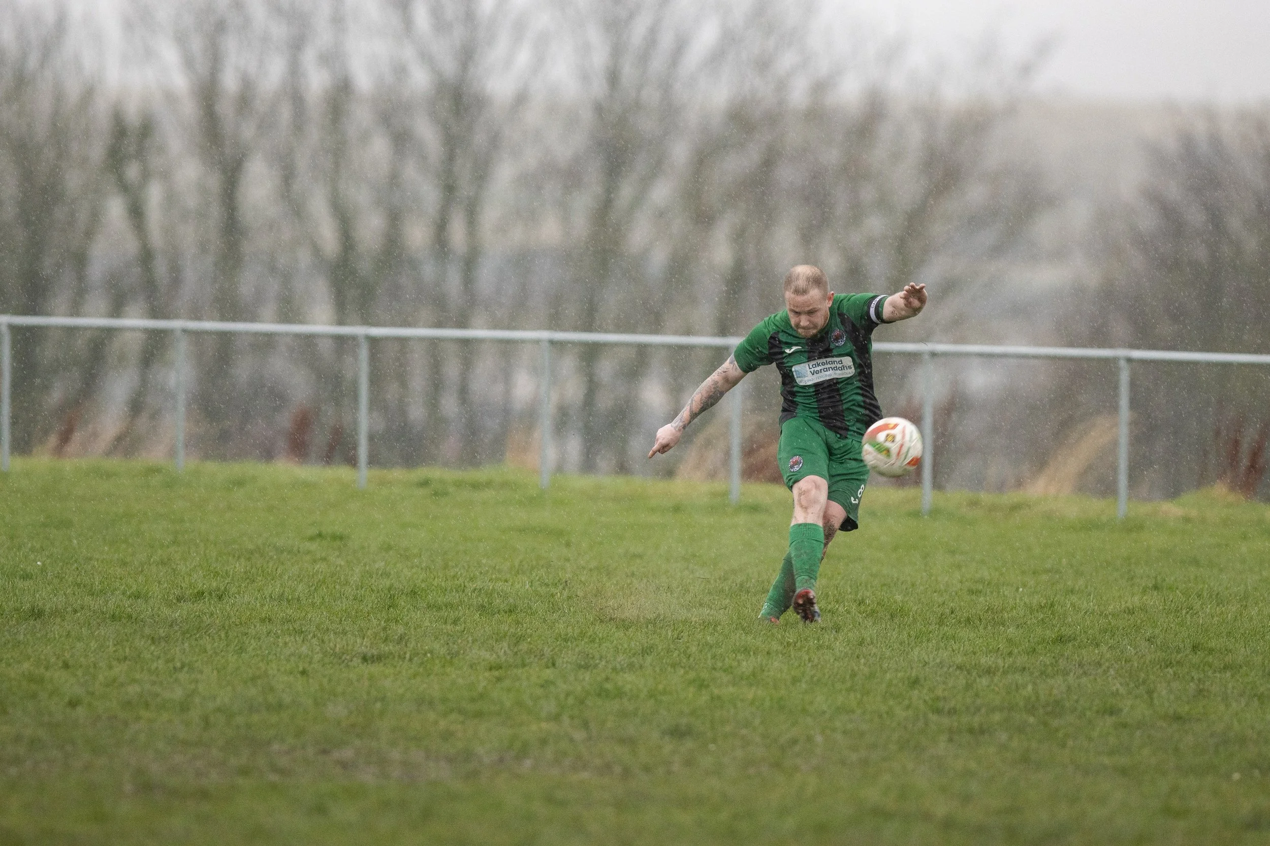 A soccer player in a green and black uniform kicking a white and colorful soccer ball on a grassy field during rainy weather.
