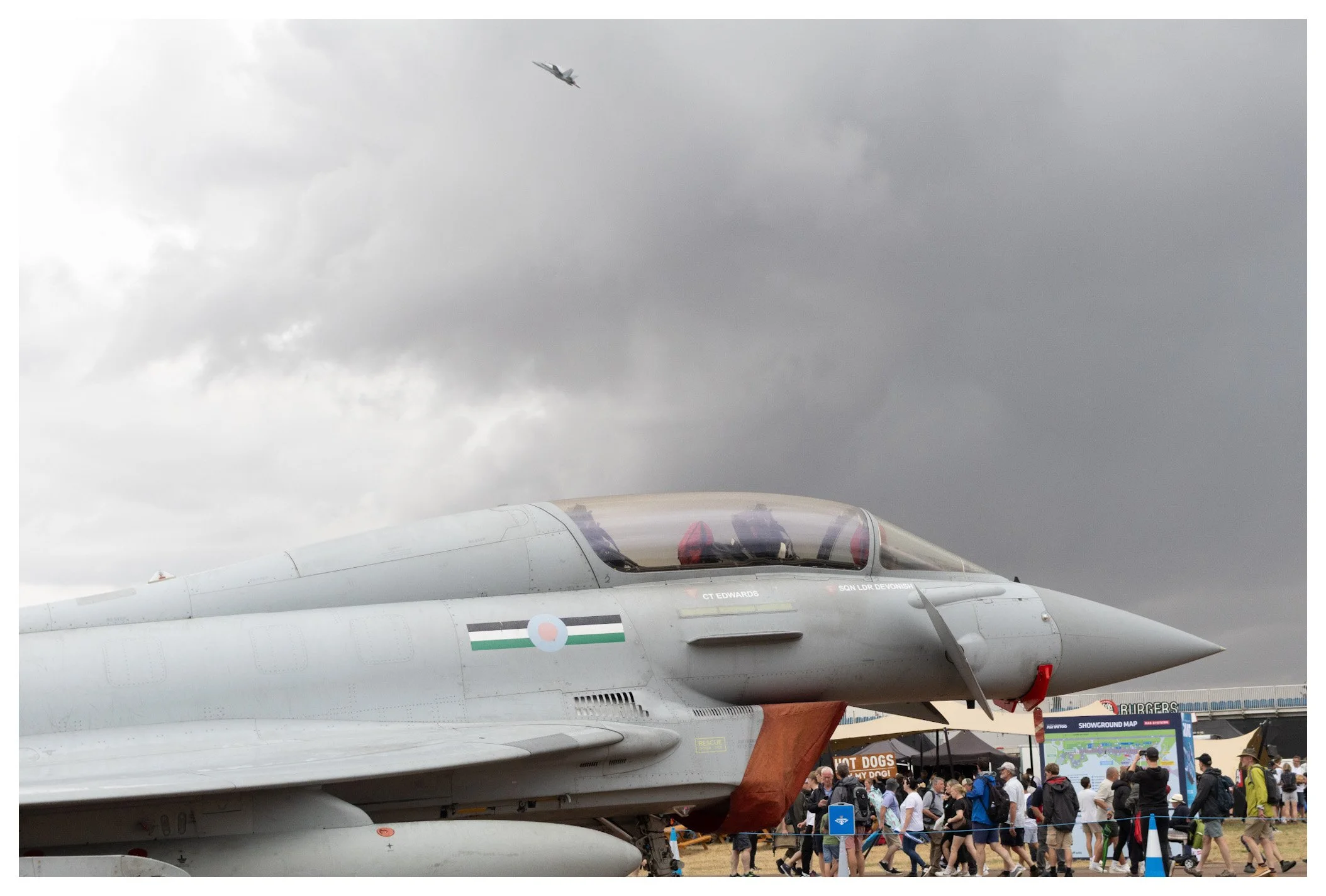 A military fighter jet on display at an outdoor event with a crowd of people walking nearby. The jet is gray with a small roundel insignia on the fuselage. There is a dark cloudy sky above, and an aircraft flying in the distance.