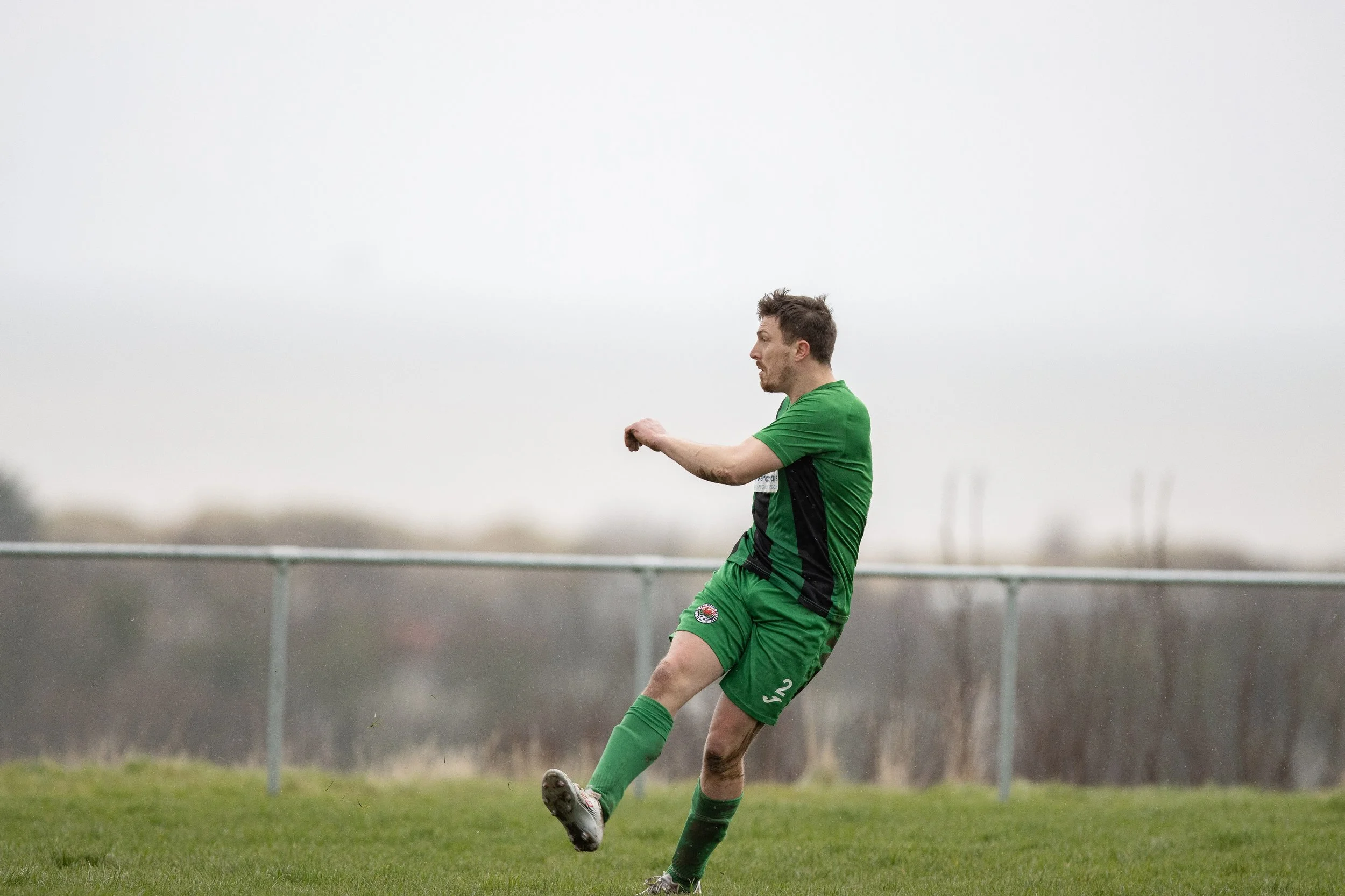 A man in a green soccer uniform kicking a ball on a grassy field with a cloudy sky in the background.