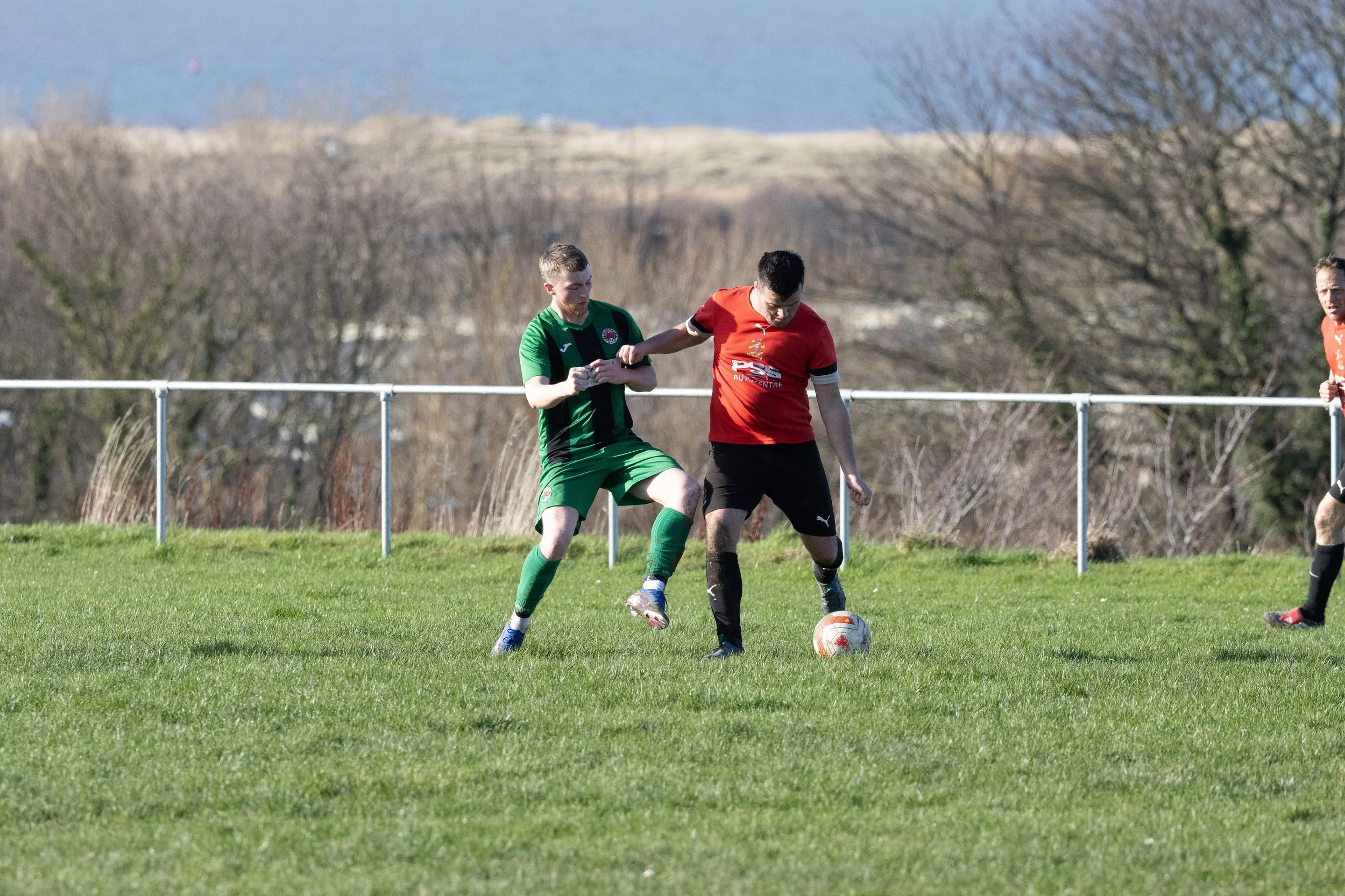 Two soccer players competing for the ball on a grassy field during a match, with a fence and leafless trees in the background.