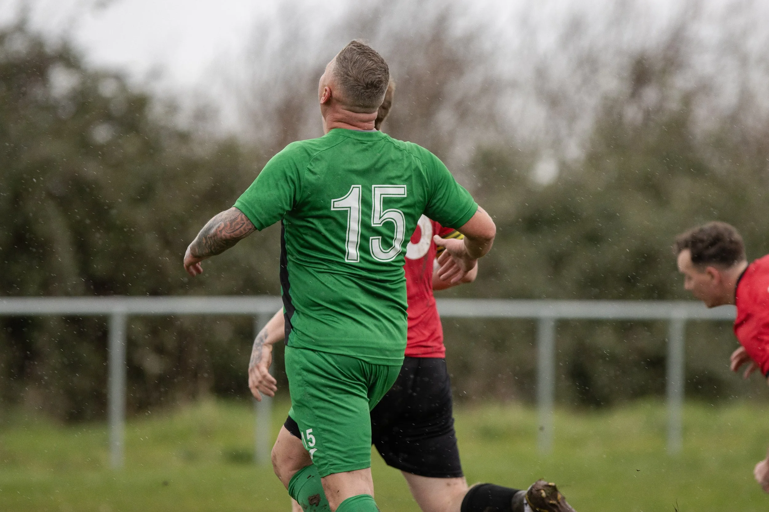 Soccer players in rain during match, one in green uniform with number 15, two others in red and black uniforms, on grassy field with trees in background.