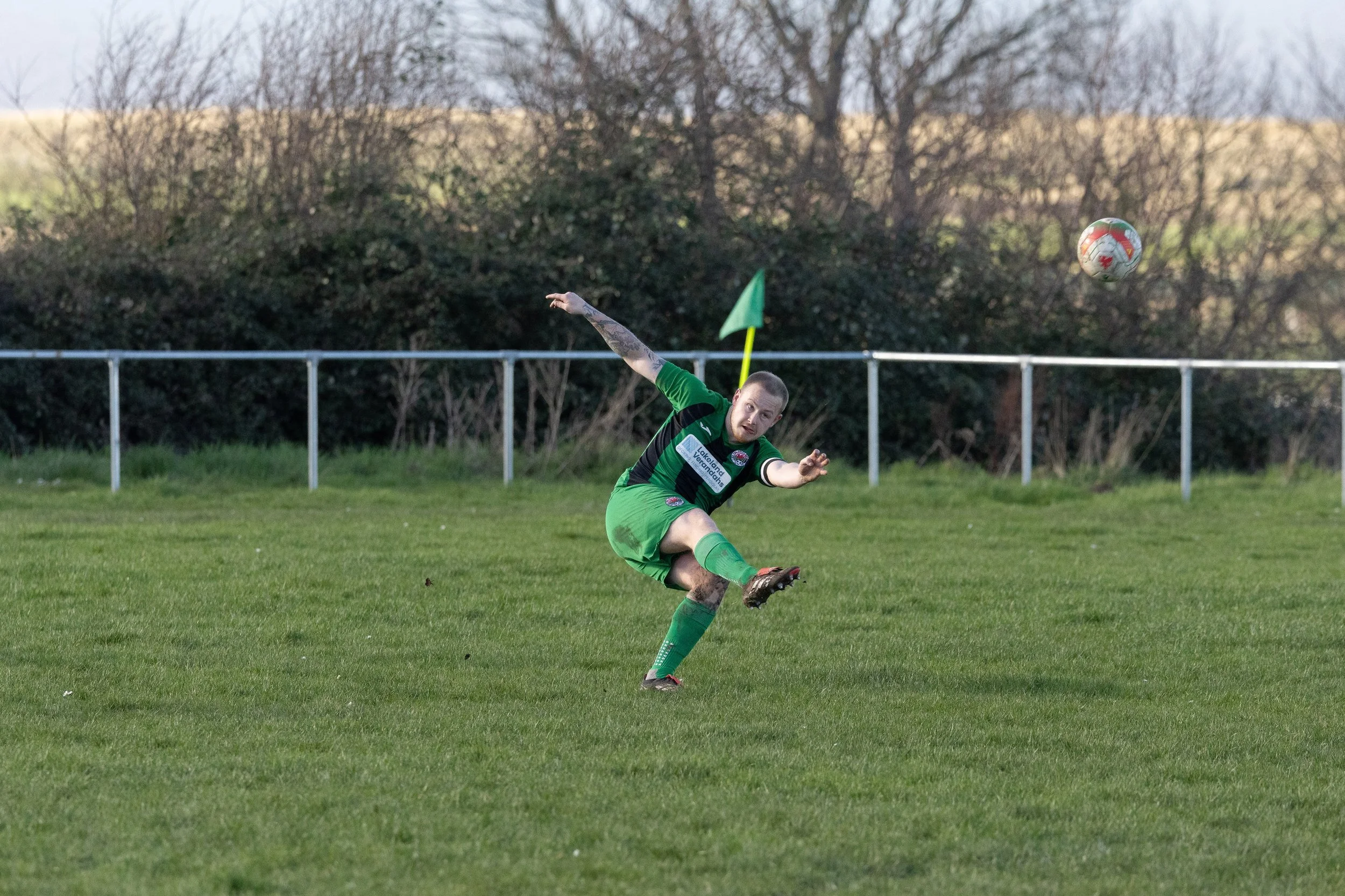 A soccer player in a green uniform is kicking a soccer ball on a grassy field during a game.