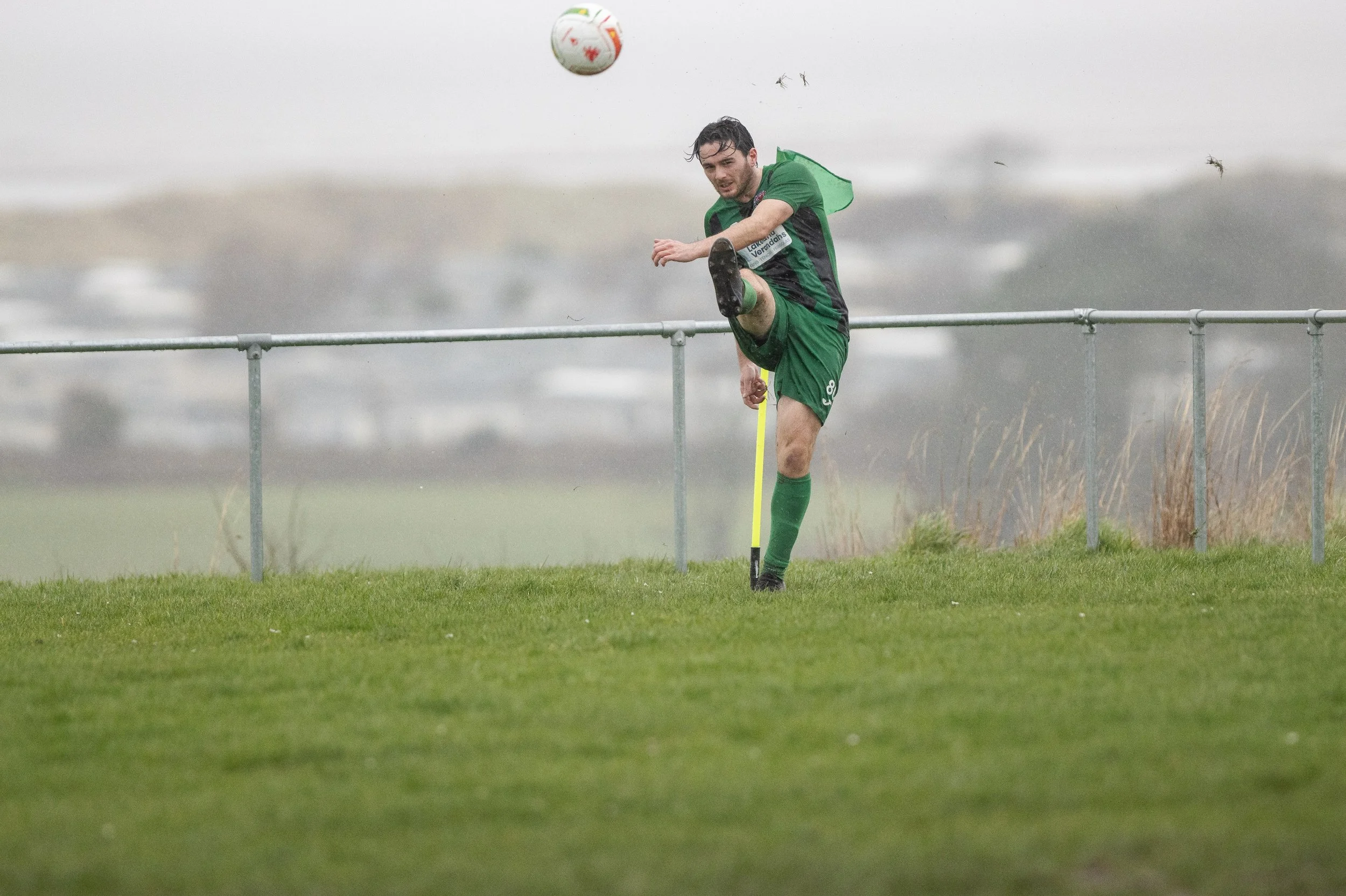 A soccer player in green uniform kicks a soccer ball on a grassy field during a game, with a foggy background and a metal railing.