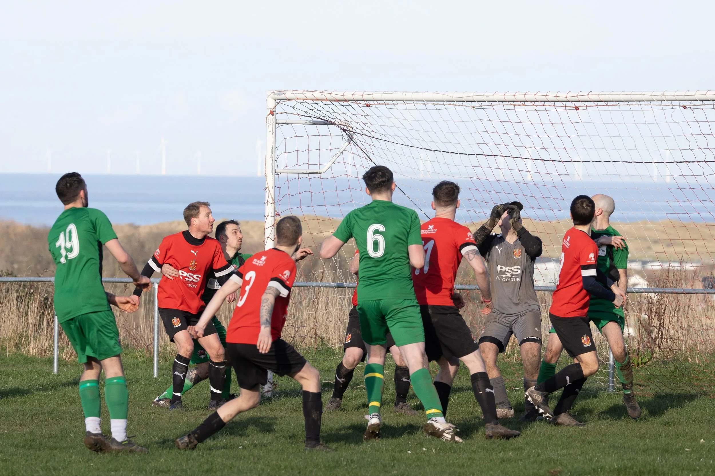 Soccer players in red and green uniforms competing near goal on grassy field with ocean in background.