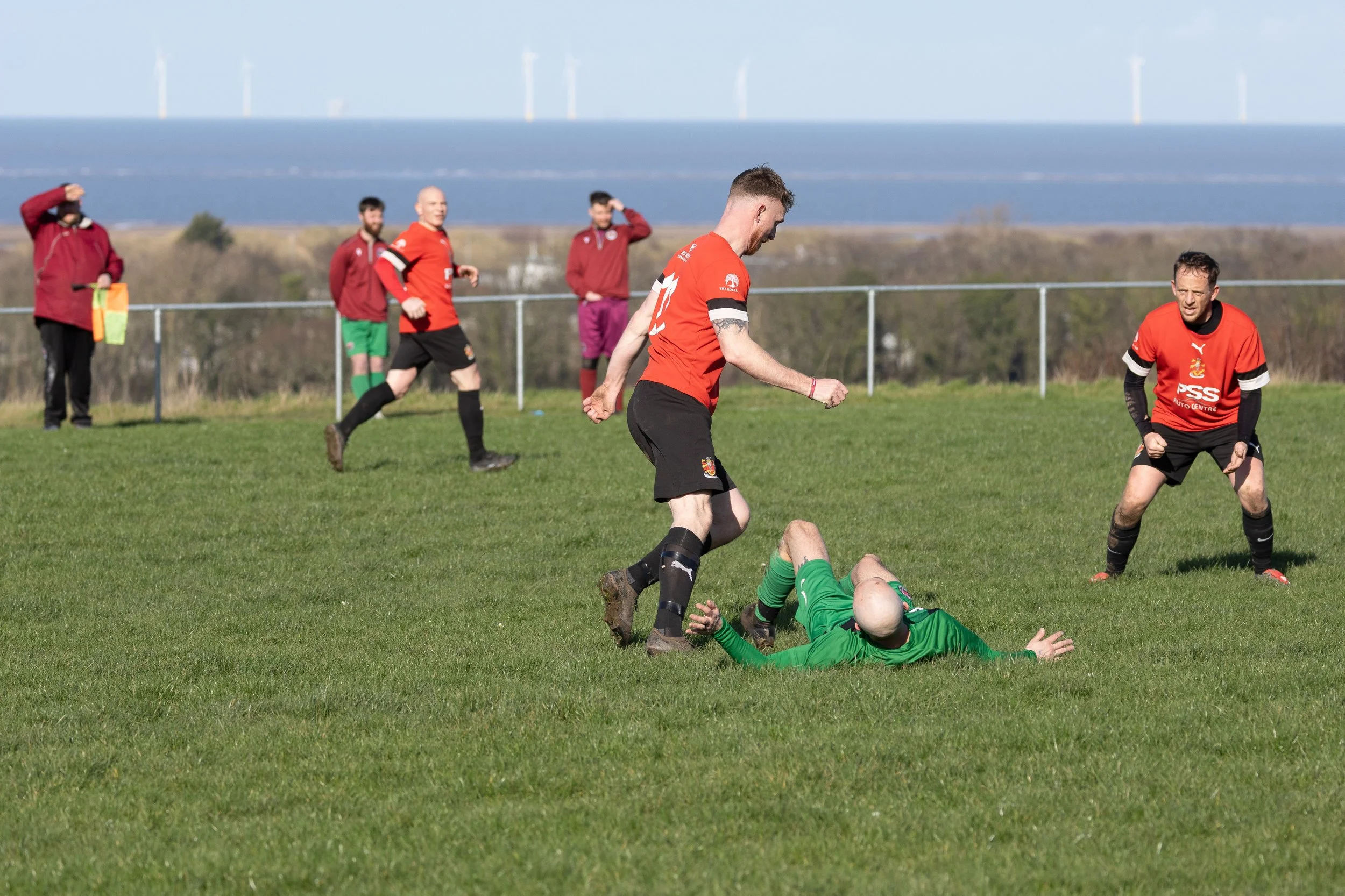 Soccer players on the field during a match, with a goalkeeper in green lying on the ground and an outfield player in red standing nearby.