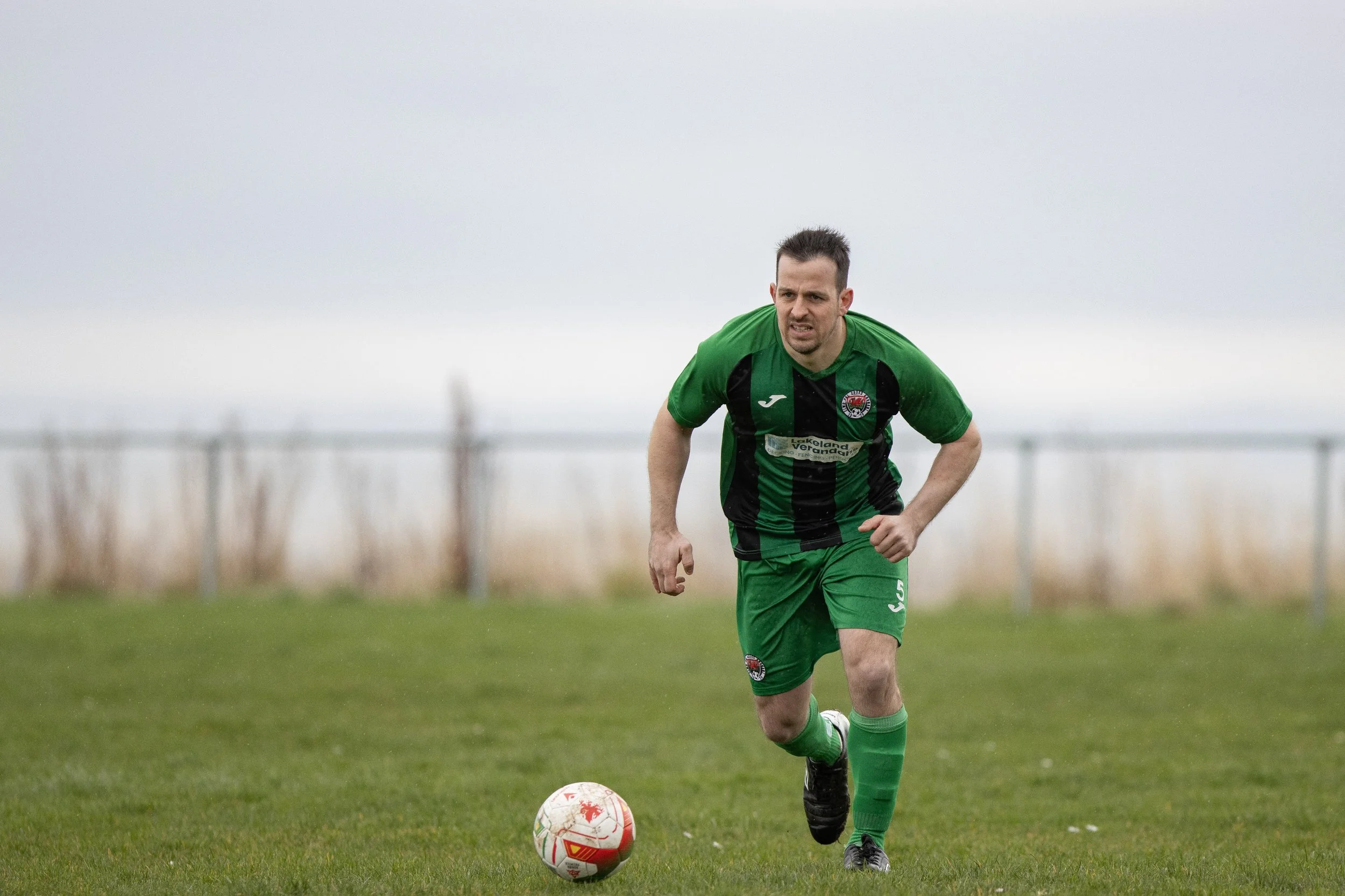 A man in a green and black soccer uniform running on a grass field, with a soccer ball in front of him, under an overcast sky.