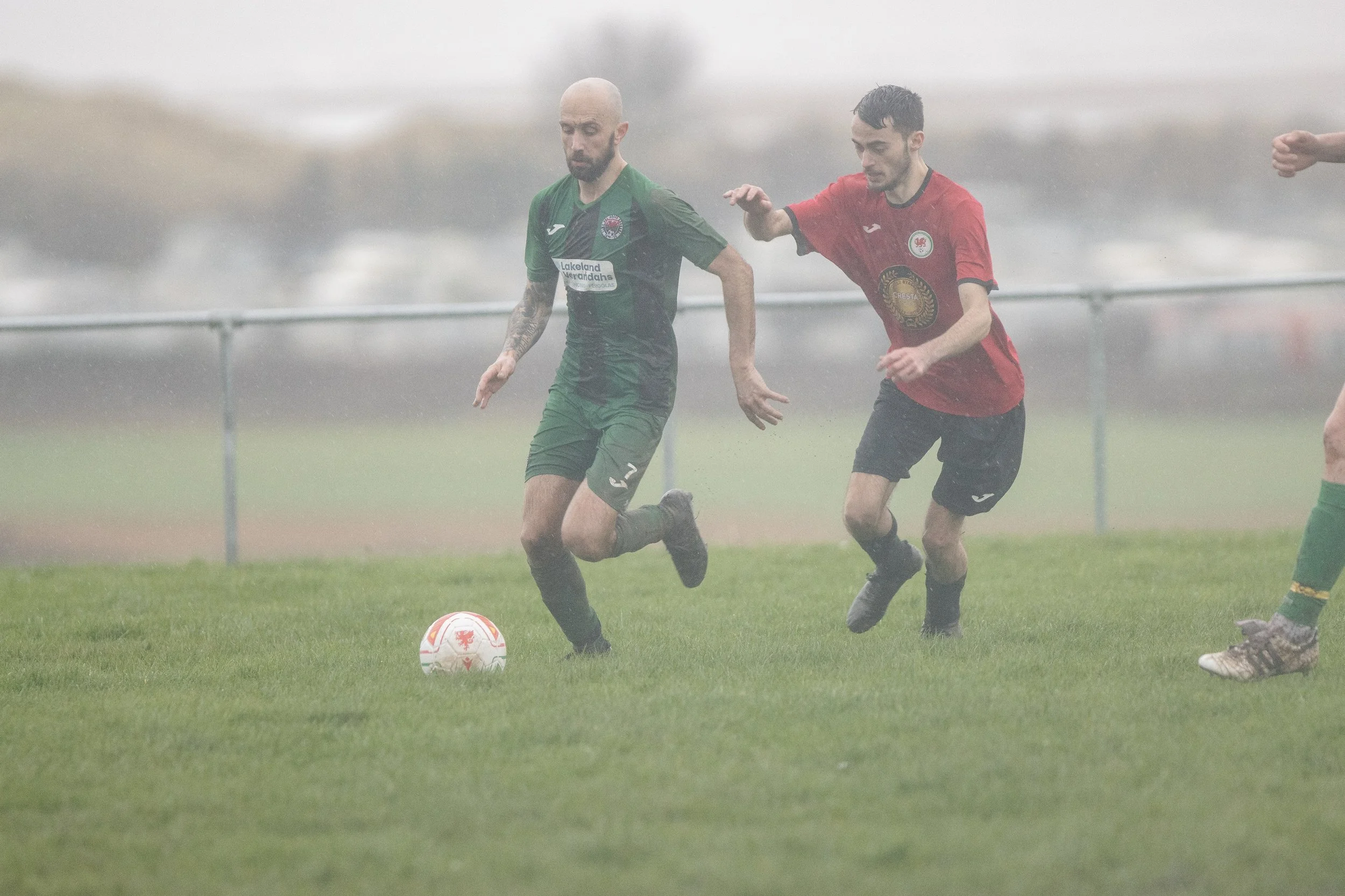 Two soccer players battling for possession of the ball on a rainy day, one in a green jersey and the other in a red jersey, on a grassy field with a fence and blurry background.