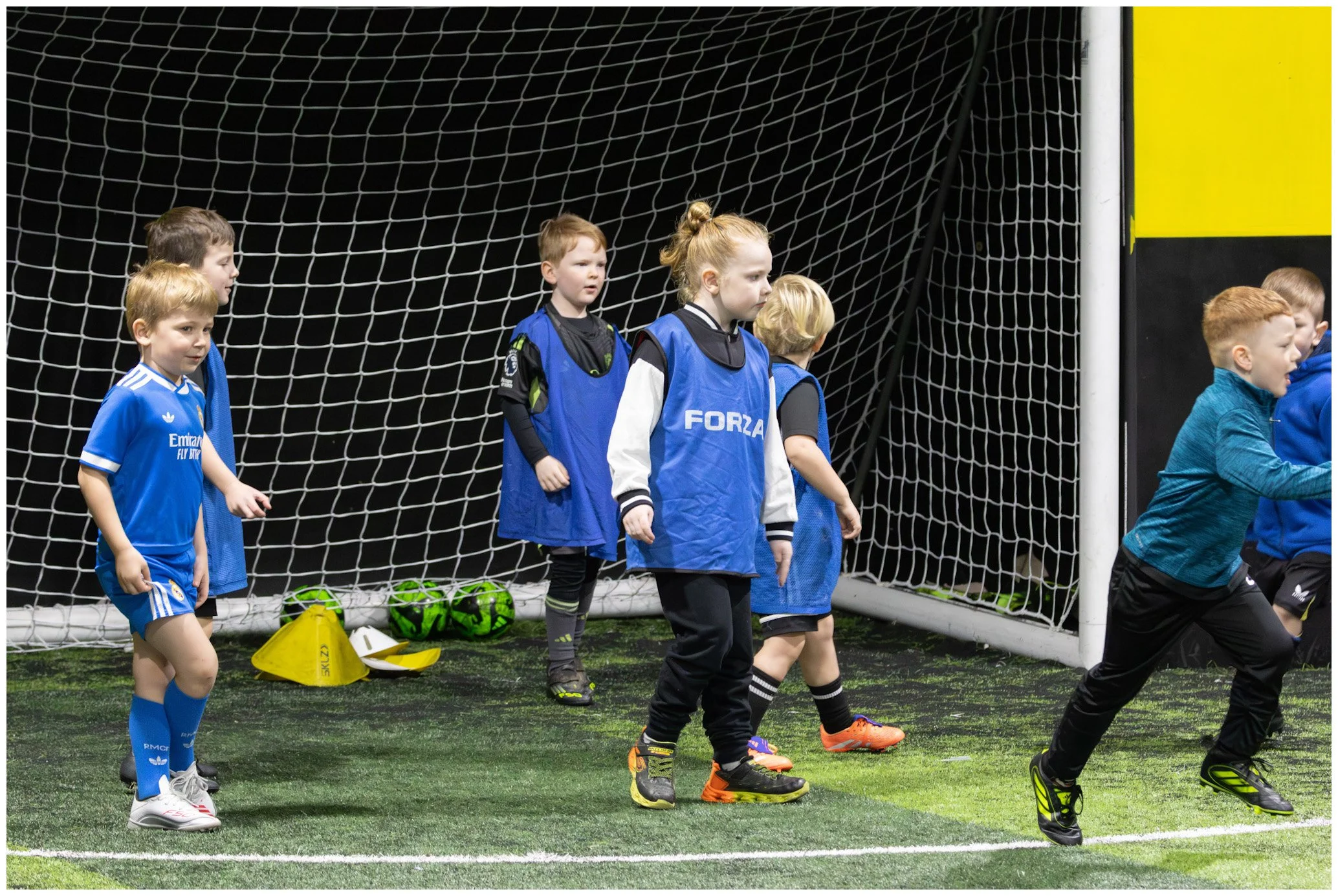 Young children participating in indoor soccer practice, standing in front of goal net, some wearing sports jerseys and training bibs.