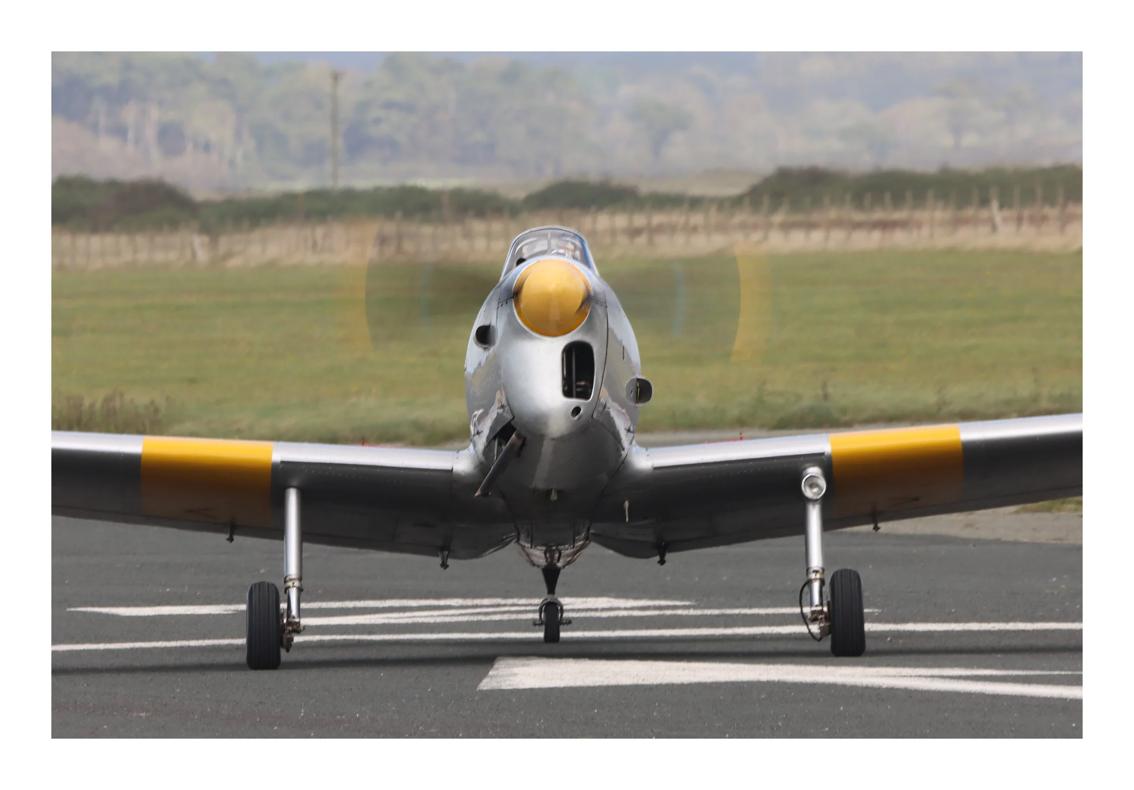 A silver military fighter jet taxiing on a runway with a blurred green field and trees in the background.