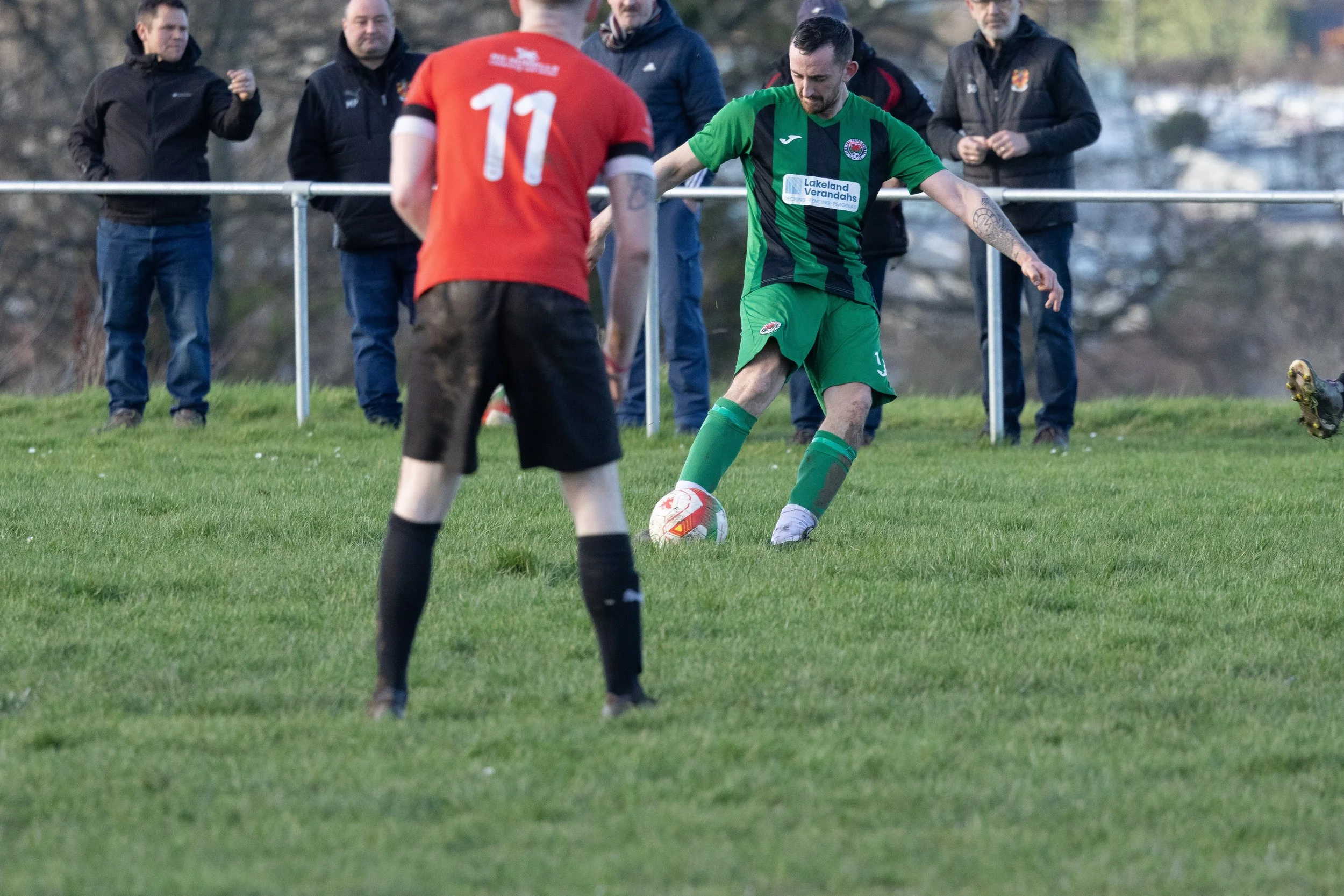 A soccer player in a green uniform is about to kick a ball during a match, with other players and spectators watching on a grassy field.