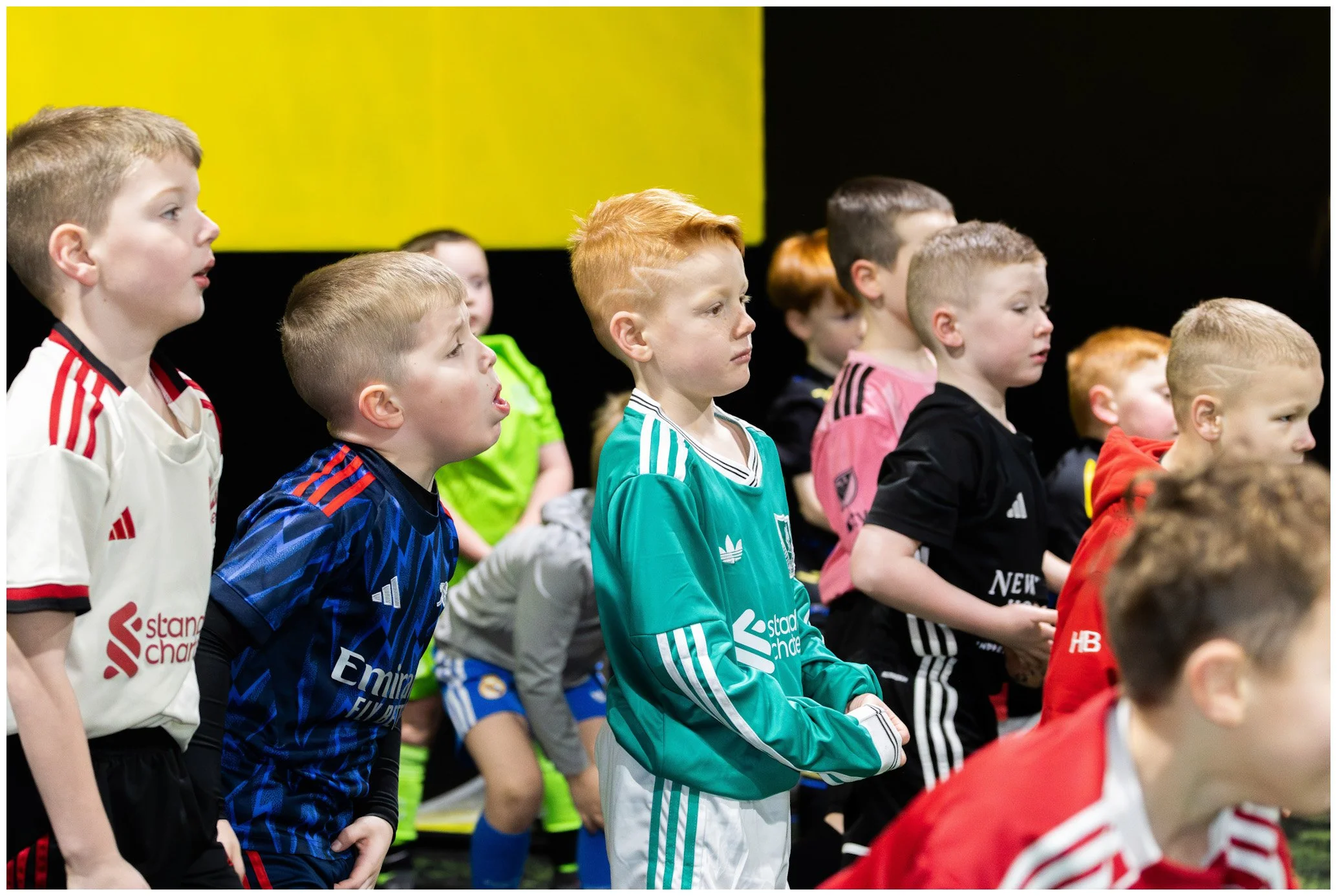 A group of young boys in colorful sports jerseys standing indoors, some with clenched fists and attentive expressions, likely watching a sports event or practicing.