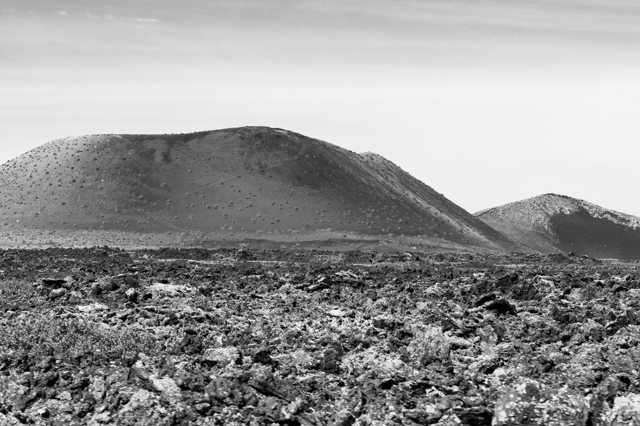 A black-and-white photo of volcanic terrain with rocky ground and two large volcanoes in the background under a partly cloudy sky.