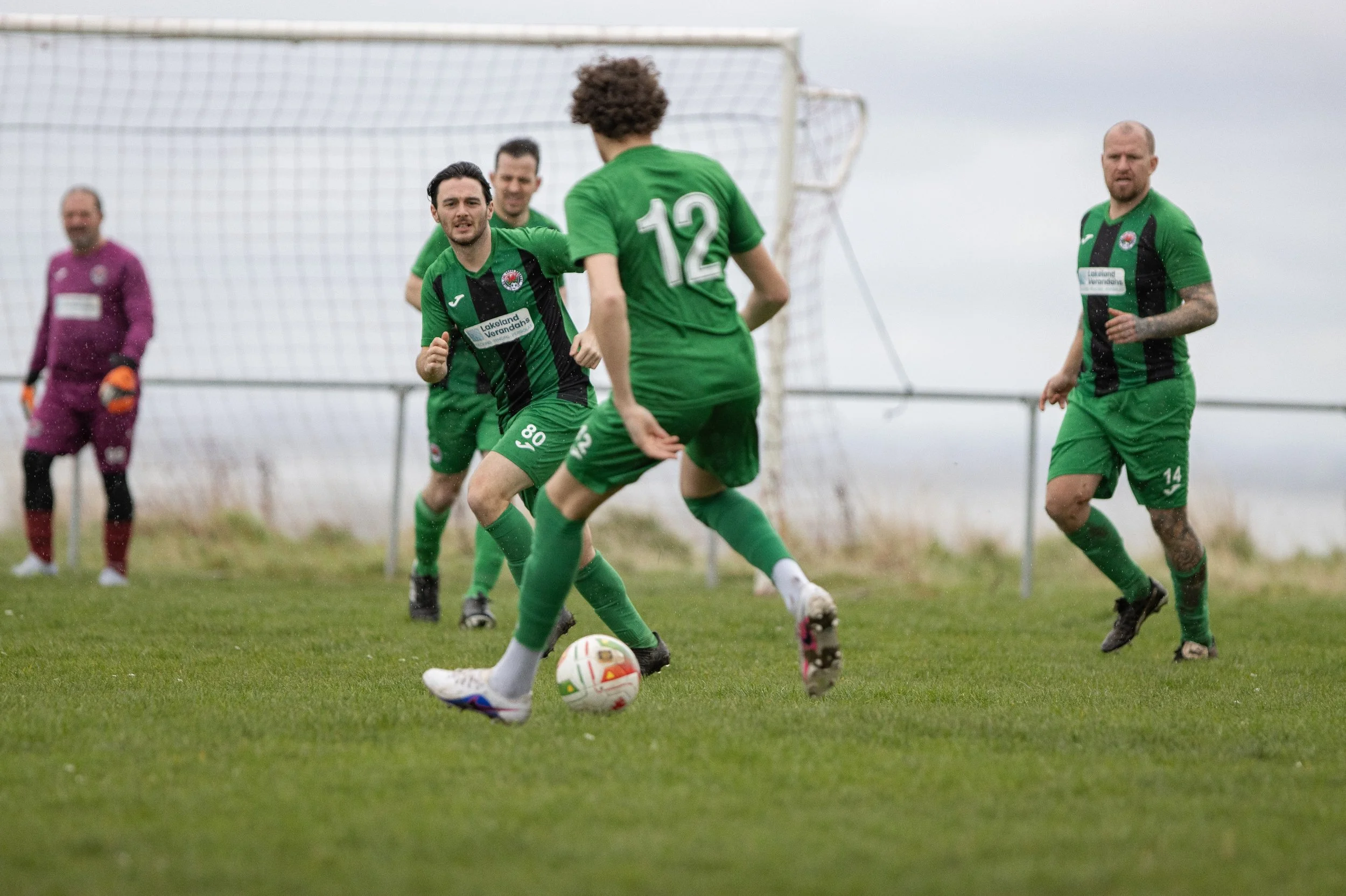 Soccer players in green uniforms on the field, with one player kicking the ball while others watch, in front of a goal under a cloudy sky.