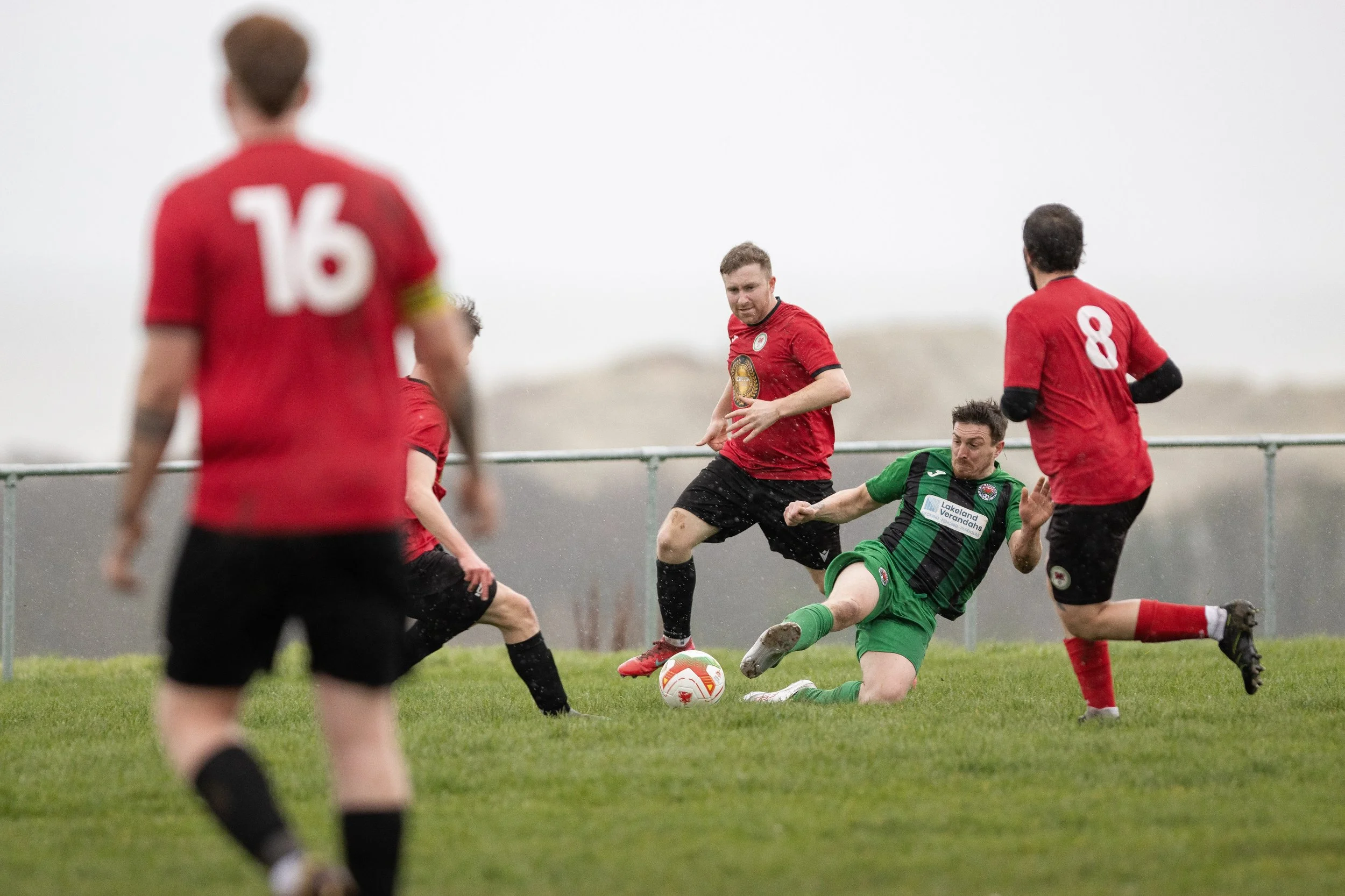 Soccer players in red and green uniforms competing for the ball on a grass field in rainy weather.