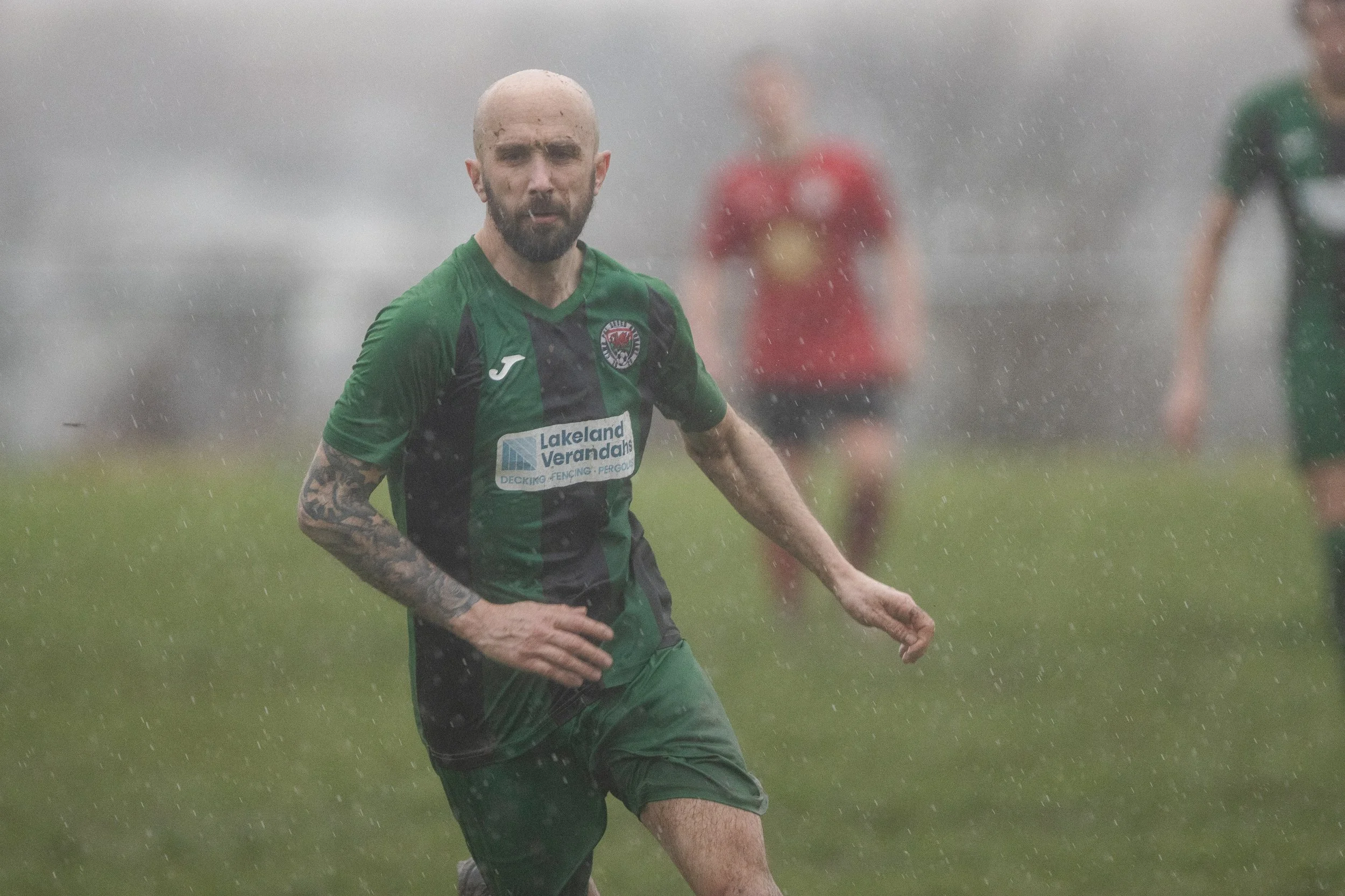 A man with a beard and tattooed arm wearing a green and black sports jersey running through rain on a grassy field, with other players in the blurred background.