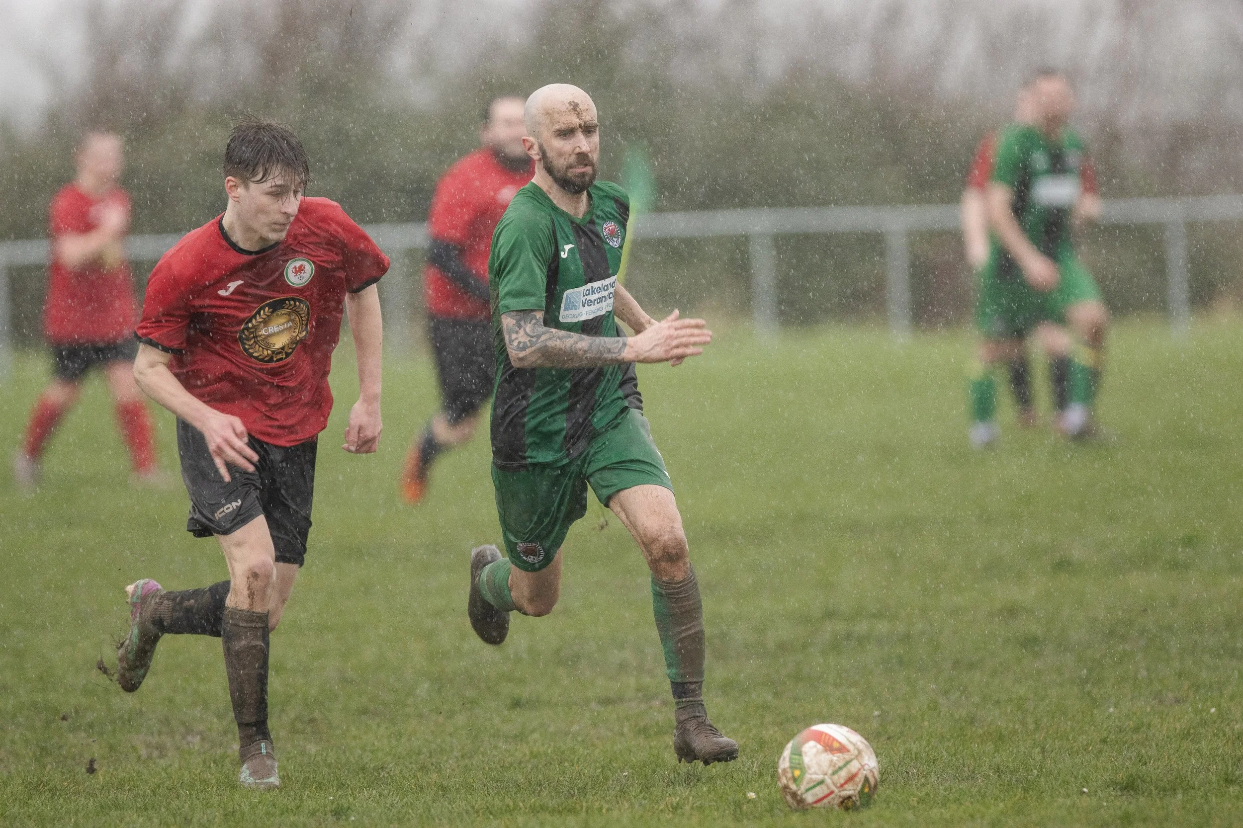 Soccer players running through a rainy field, one in a green and black uniform and another in a red uniform, with a soccer ball in front of them.