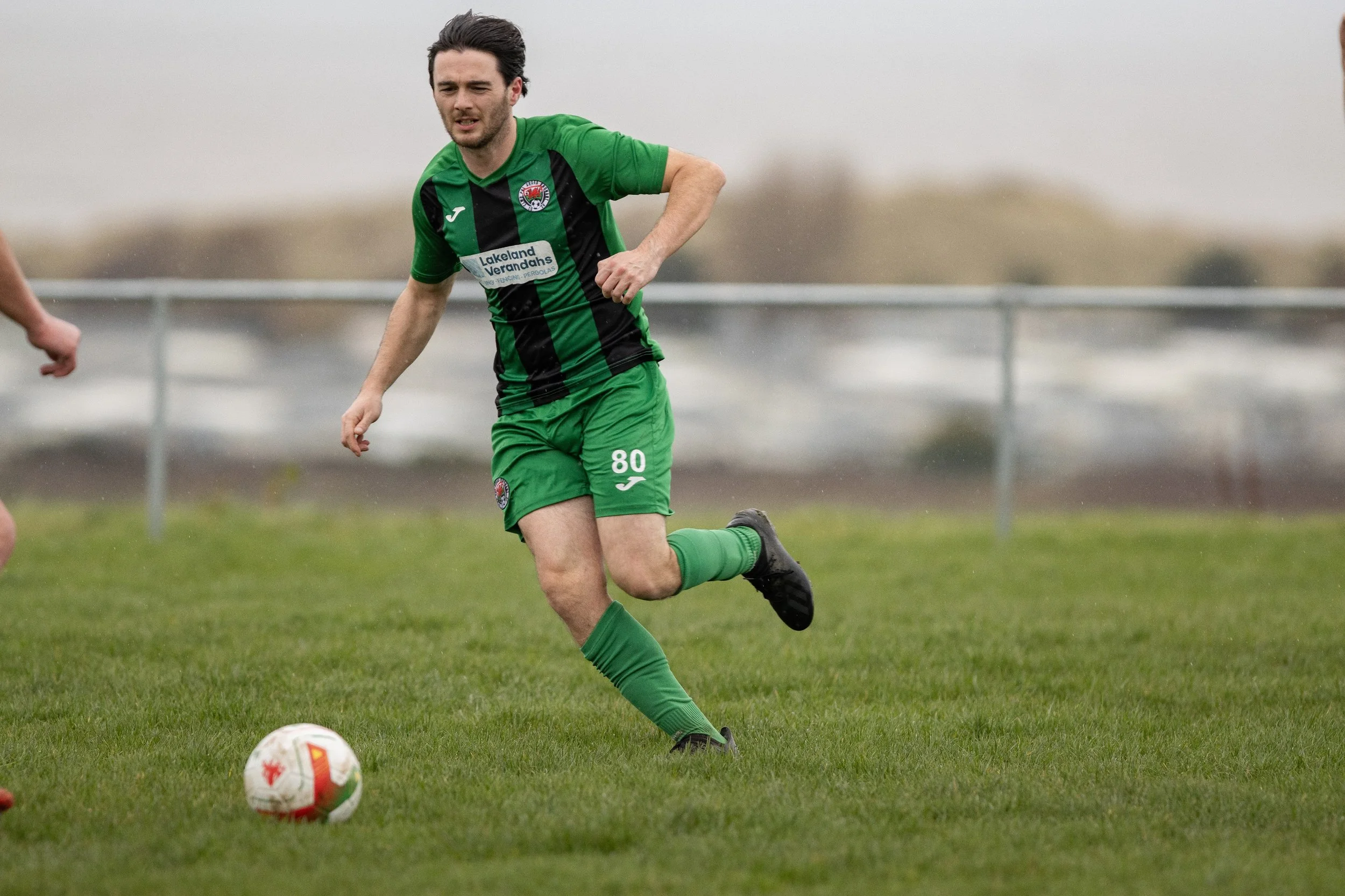 A male soccer player in green and black uniform running on a grassy field with a soccer ball near his feet.