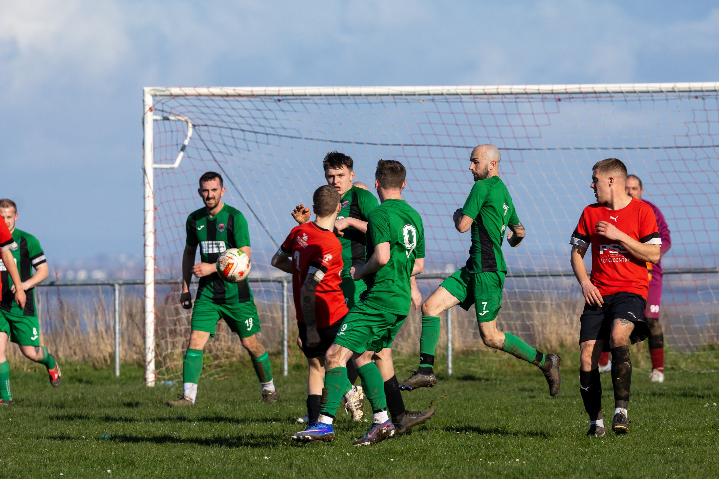 Soccer players in green and red uniforms compete for the ball near the goal on a grassy field with a blue sky background.
