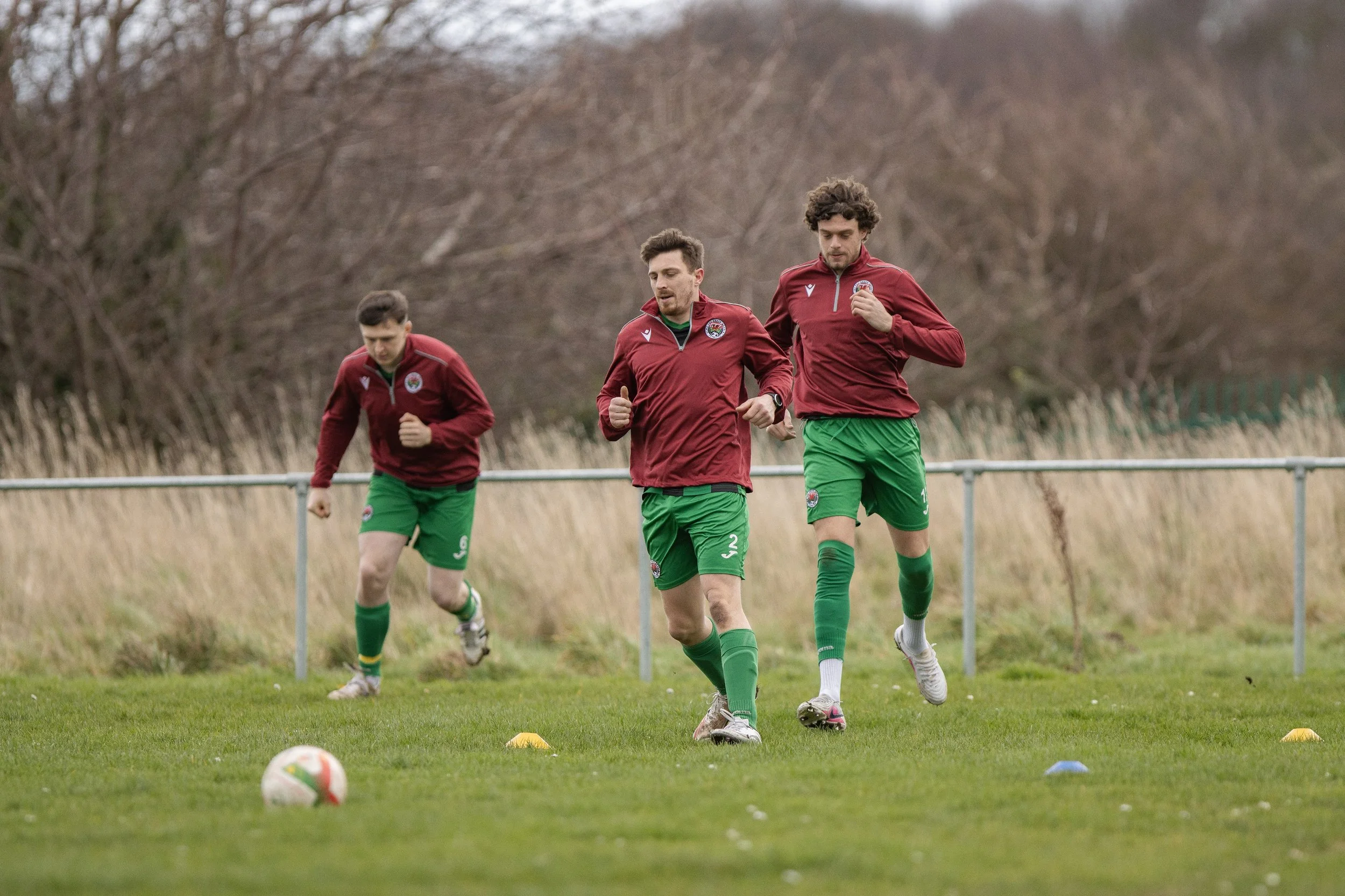 Three soccer players in red jerseys and green shorts jogging on a grassy field during practice, with orange and blue training cones on the ground and a background of bare trees.