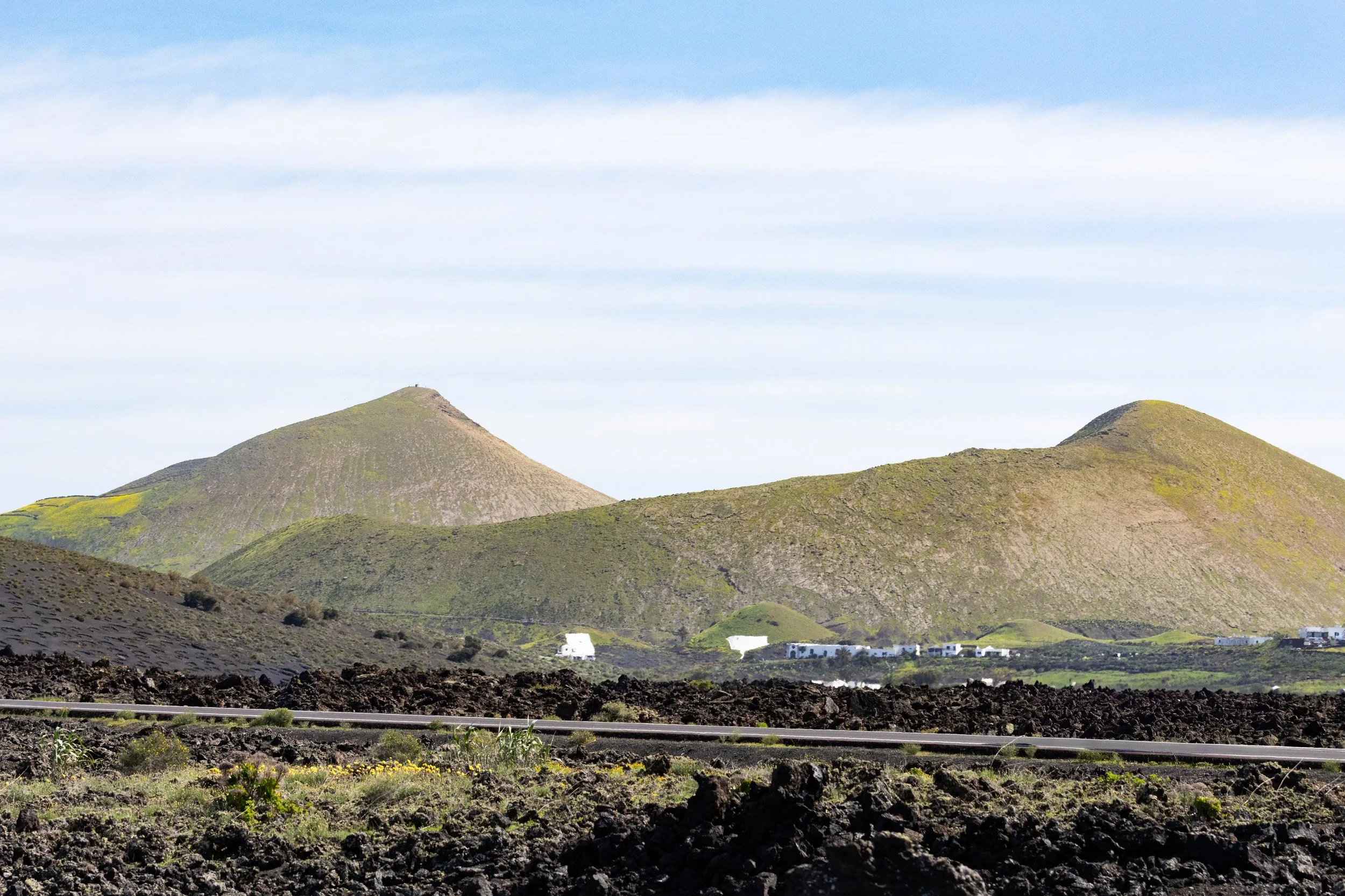 Scenic view of lush green volcanic hills with a partly cloudy sky in the background, arid foreground with volcanic rocks, and some white buildings at the base of the hills.