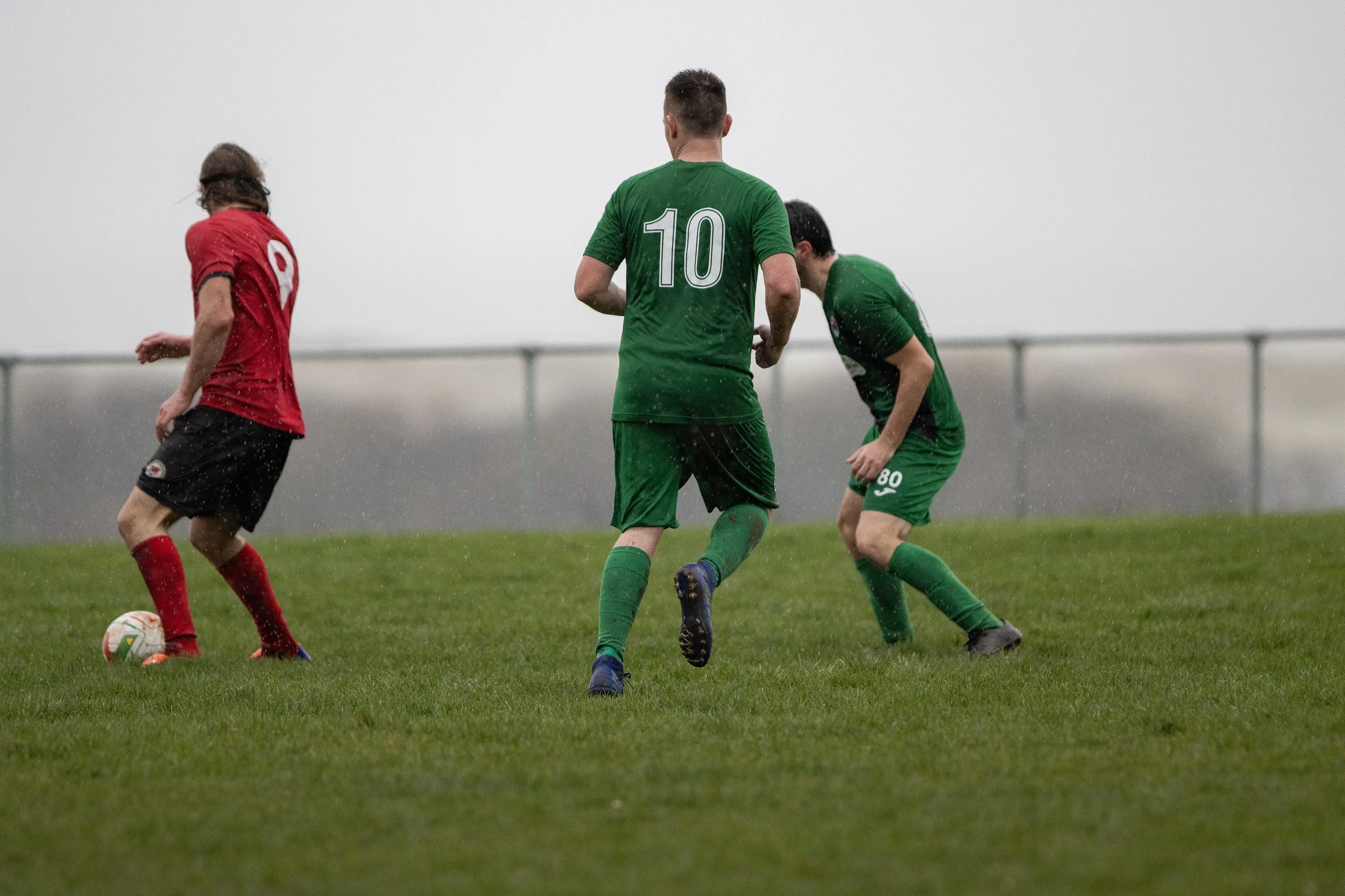 Three soccer players on a rain-soaked field, one wearing a red jersey with the number 9, and two wearing green jerseys with the numbers 10 and 80, during a match in rainy weather.