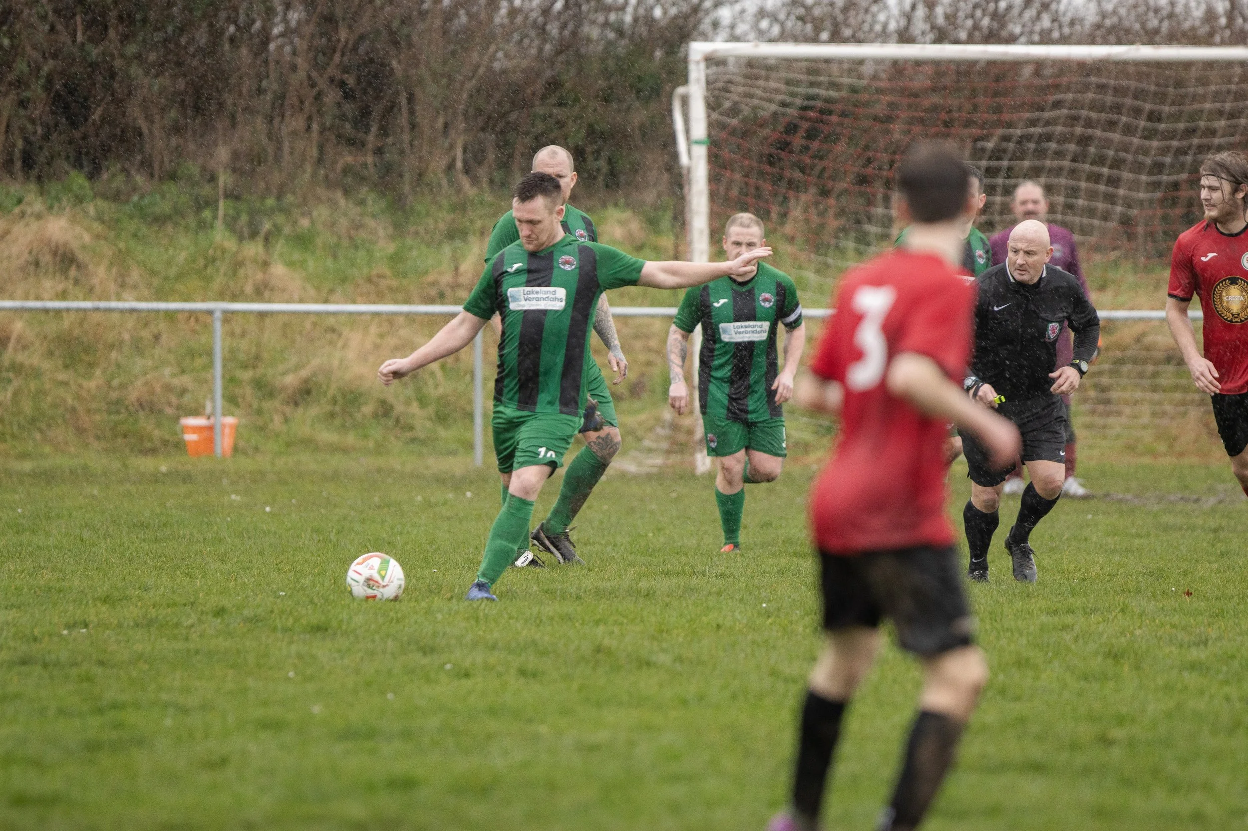 Soccer match with players in green and red jerseys on a muddy field during rainy weather, with a goal net in the background.