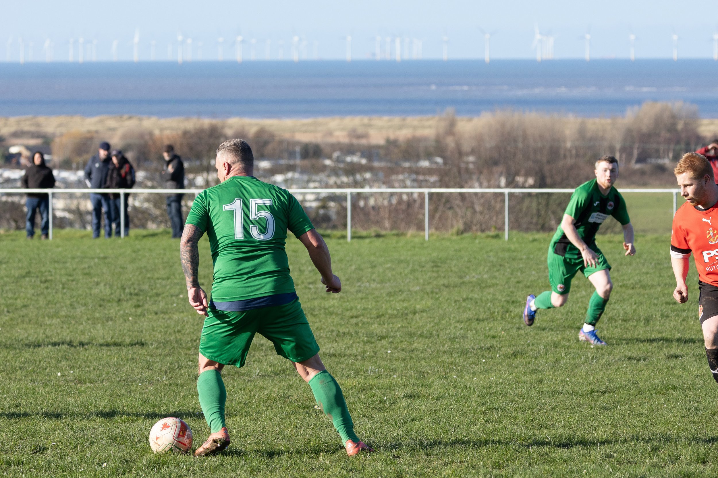 Soccer players on a grassy field, one in a green jersey with the number 15, preparing to kick the ball, with a scenic backdrop of the ocean and wind turbines in the distance.